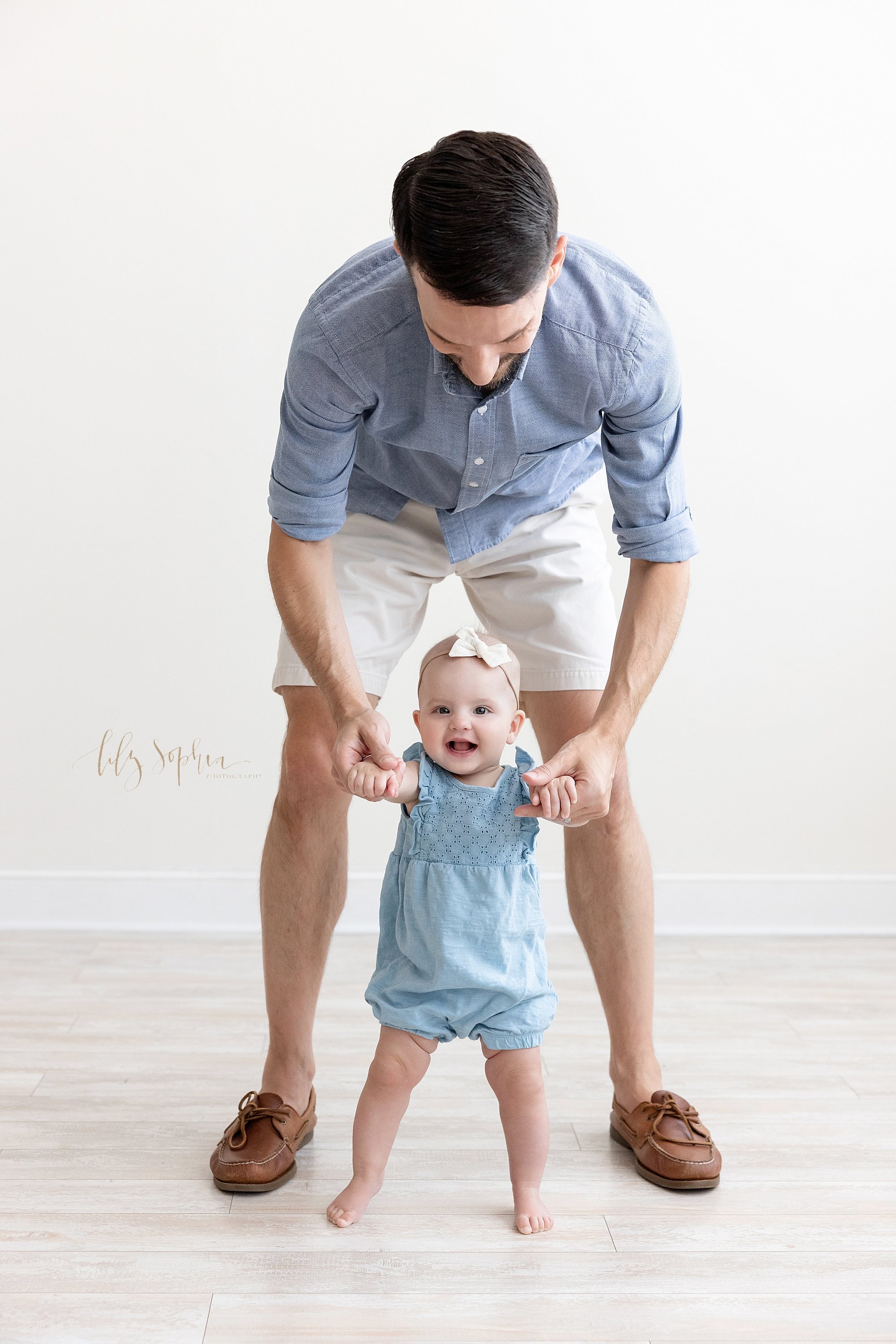  Family photo shoot of a little girl as she holds onto her father’s hands and walks in front of him taken in a studio near Buckhead in Atlanta that uses natural light. 