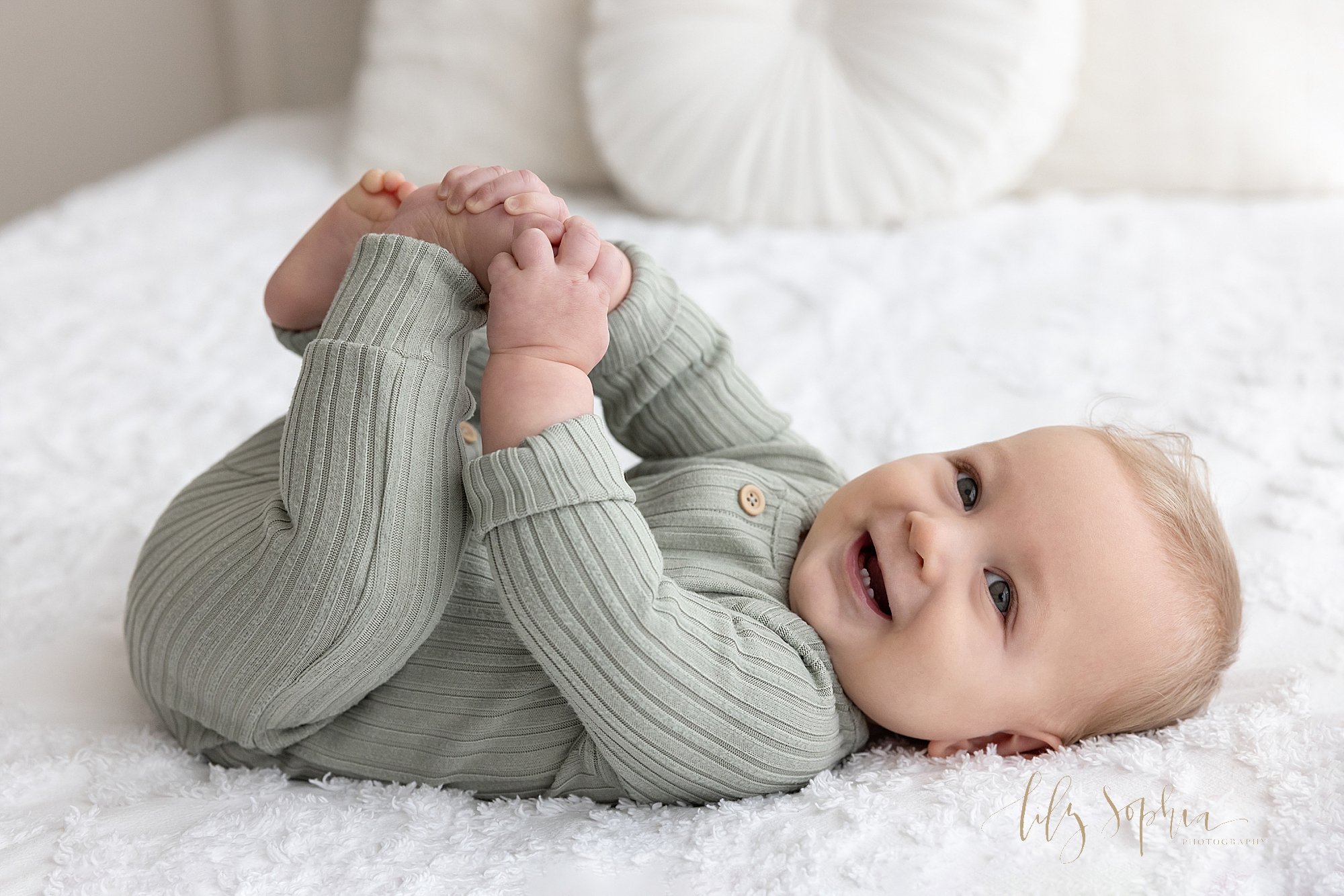  Baby portrait of a baby boy lying on his back on a bed as he smiles to show his tiny bottom teeth and plays with his toes taken near Virginia Highlands in Atlanta, Georgia in a studio that uses natural light. 