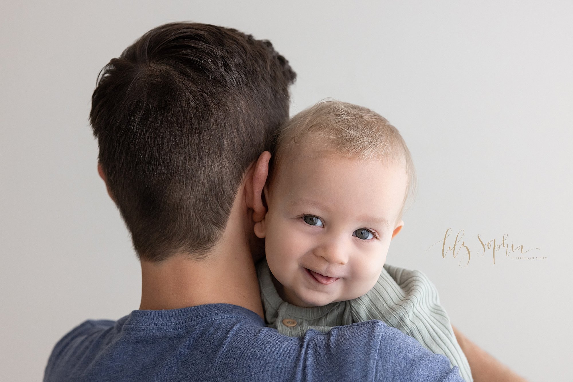  Baby picture of a baby boy sticking out his tongue as he looks over his father’s shoulder as he holds him taken near Poncey Highlands in Atlanta in a photography studio that uses natural light. 