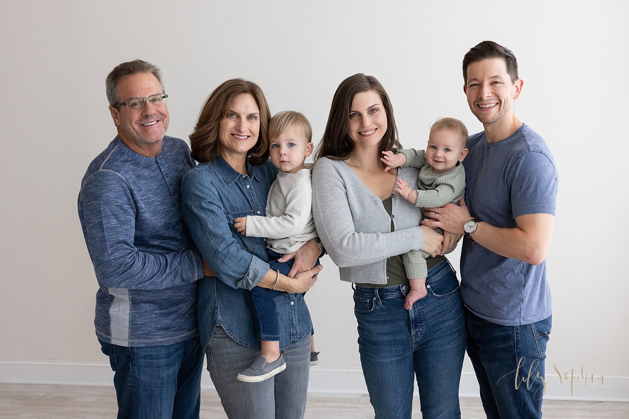  Multi-generational family portrait of a grandfather standing next to his wife who is holding their toddler grandson in her arms as she stands next to her daughter who is holding her baby boy in her harms as her husband stands beside her taken using 