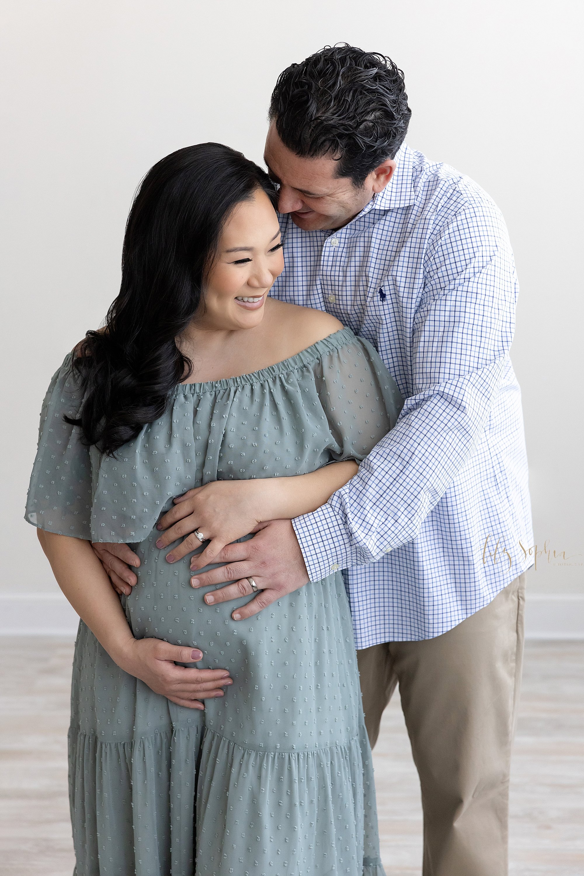  Maternity shoot with an expectant couple as the husband stands behind his wife and wraps his arms around her to place his hands on her belly while his wife turns her head to the right while framing her belly with her hands and her husbands whispers 