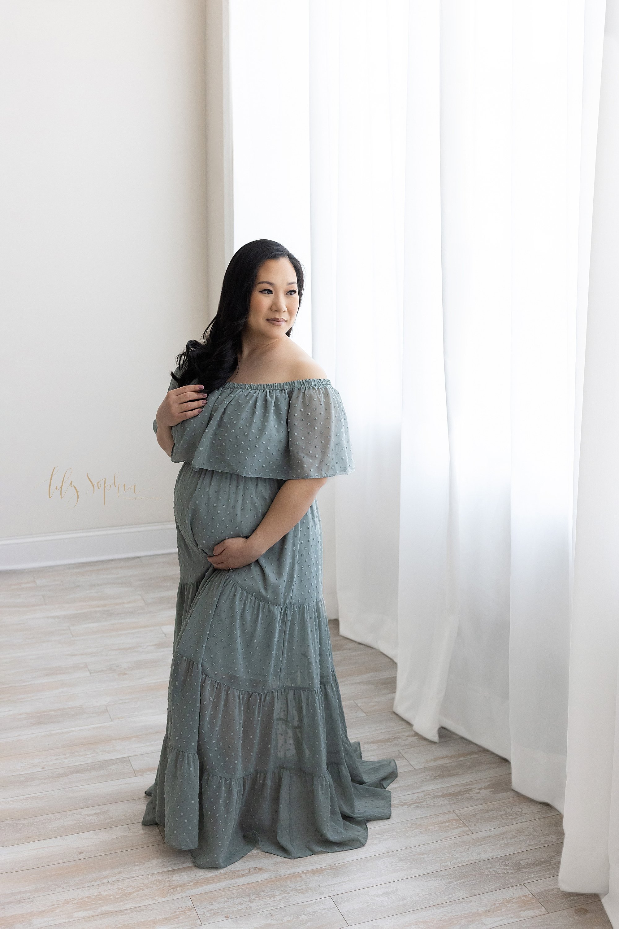  Maternity portrait of a pregnant mother wearing an off the shoulders flocked gown with a large bodice as she stands looking over her left shoulder while playing with her hair with her right hand and holding the base of her belly with her left hand i