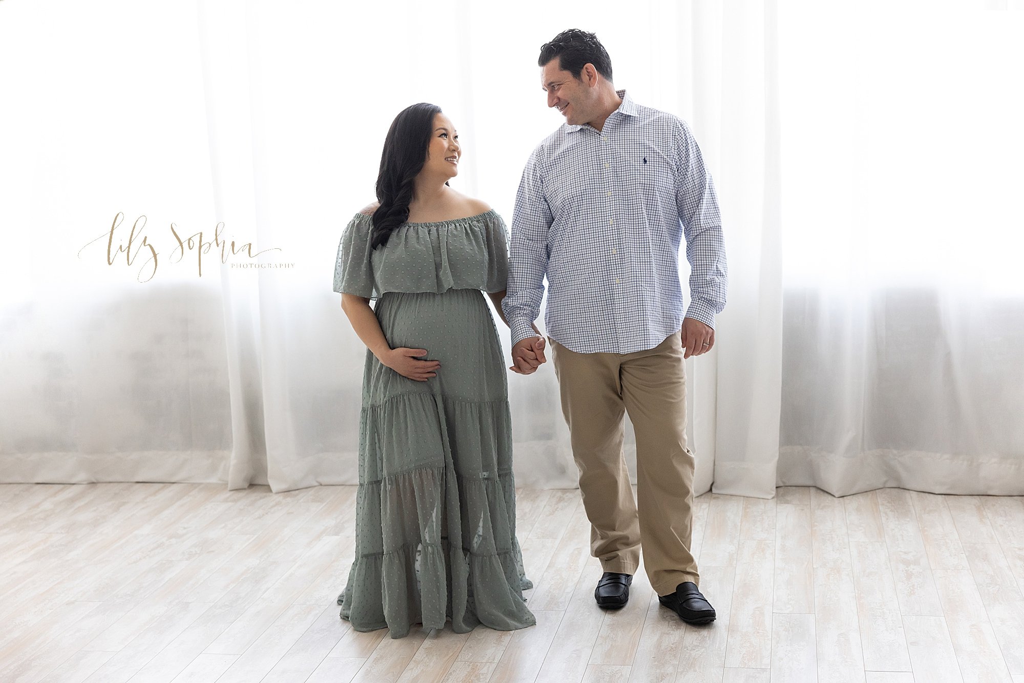  Maternity picture of a husband holding his pregnant' wife’s hand as they look at one another and mom holds her belly taken near Kirkwood in Atlanta in front of a window streaming natural light in a photography studio. 