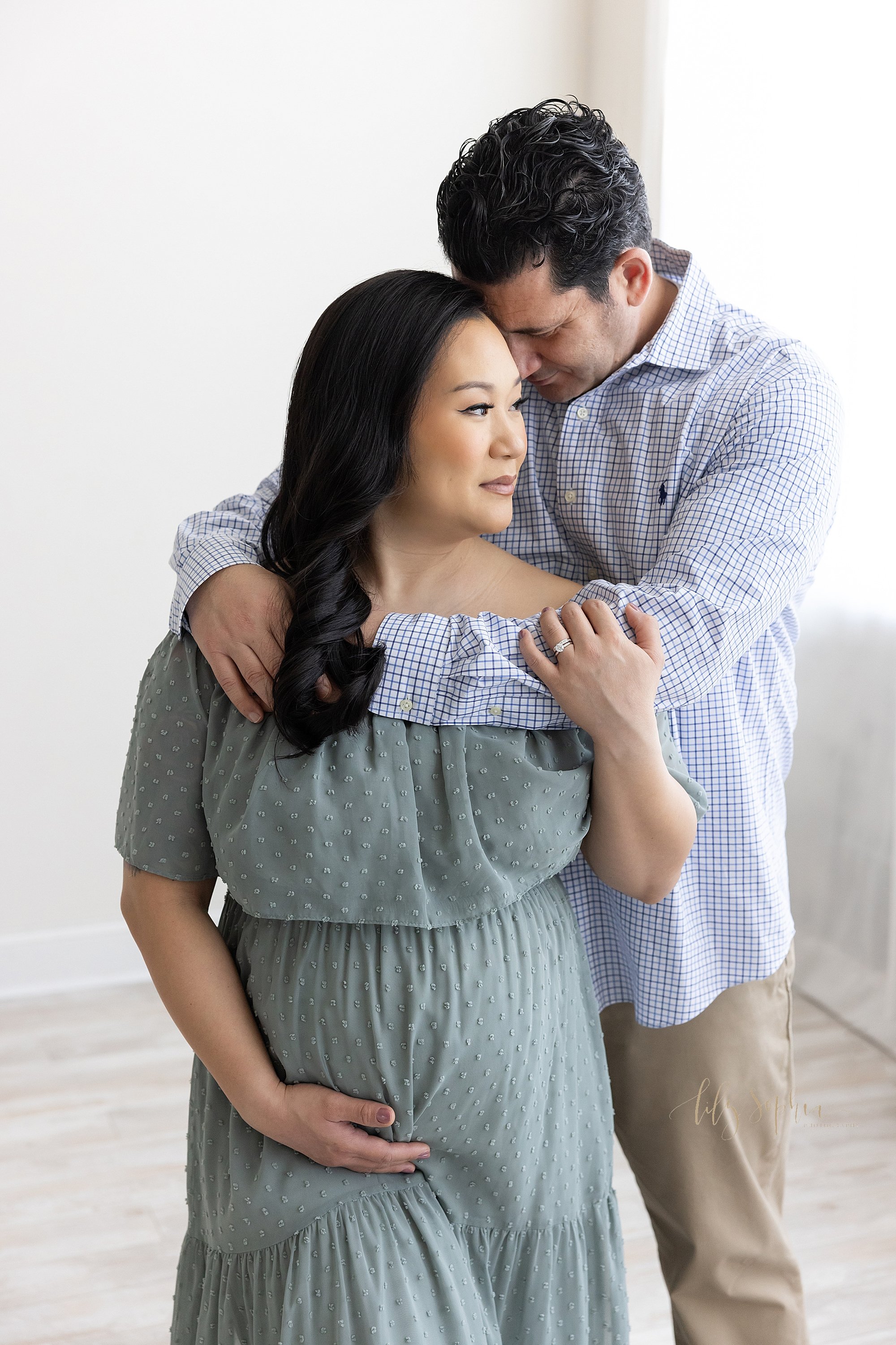  Maternity portrait of a husband standing behind his wife and wrapping his arms around her shoulders as he places his forehead on her head while she looks over her left shoulder, holds his arm with her left hand and holds the base of her belly with h