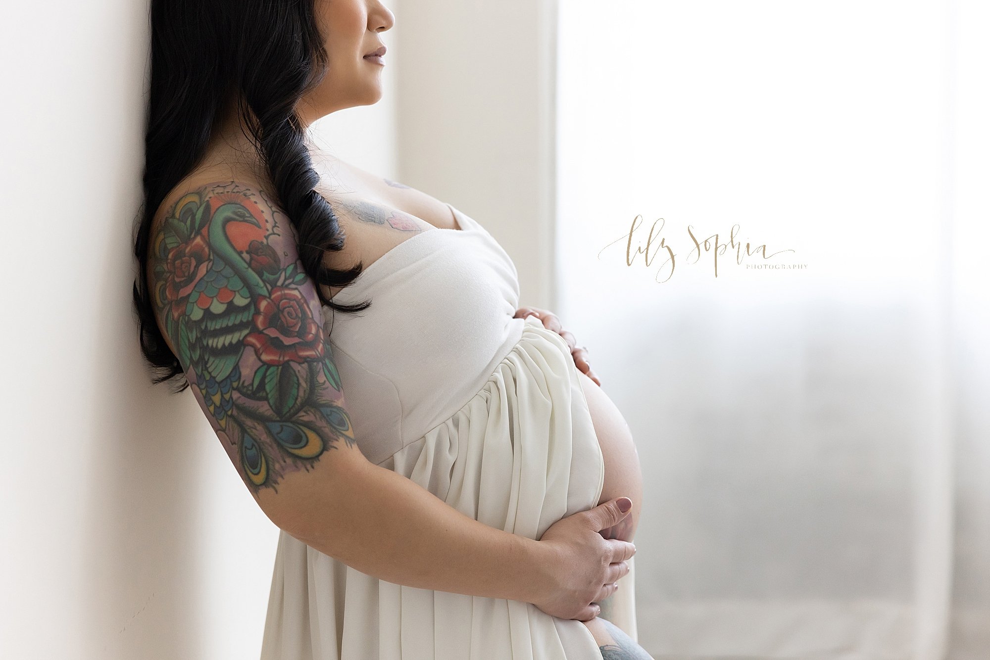  Maternity session with an Asian woman wearing a strapless split front gown as she stands with her back to a ball and framing her belly with her hands next to a window streaming natural light into a photography studio located near Midtown in Atlanta,