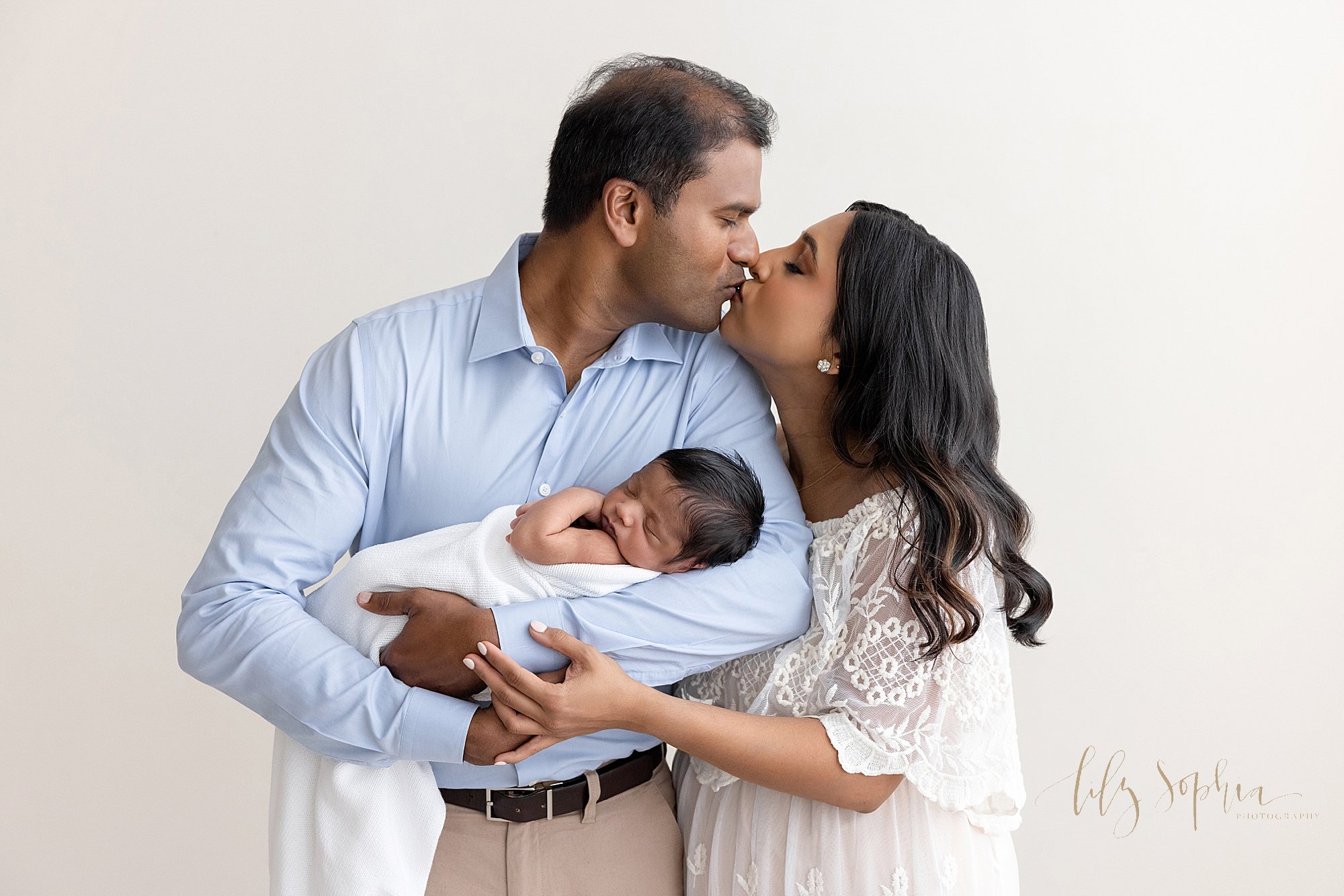  Family newborn photo session with a father cradling his sleeping newborn baby boy in his arms as he turns his head over his left shoulder to kiss his wife who is standing next to him taken in natural light in a studio near Virginia Highlands in Atla
