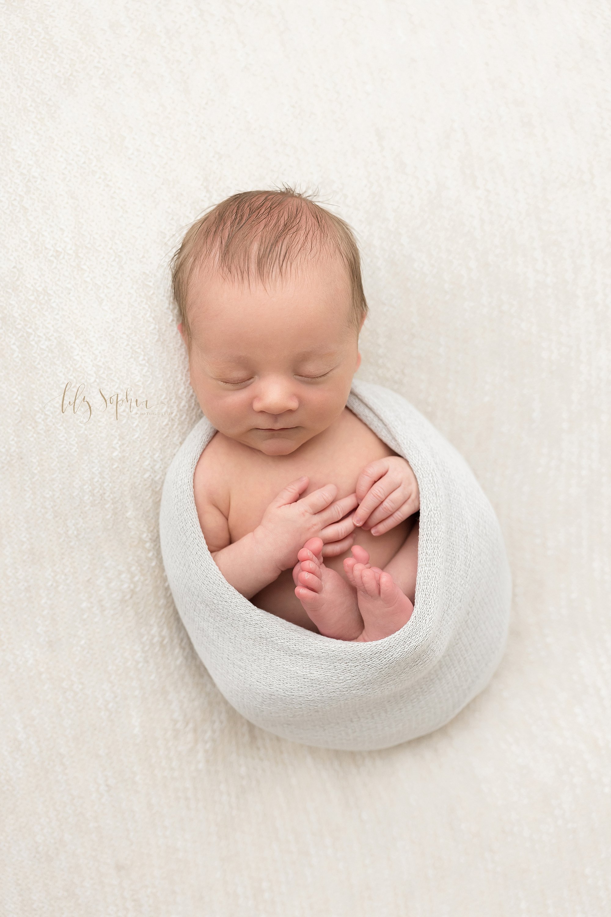  Overhead newborn picture of a newborn baby boy cradled in a stretchy swaddle with his delicate fingers and his feet sticking out taken in a studio near Morningside in Atlanta, Georgia that uses natural light. 