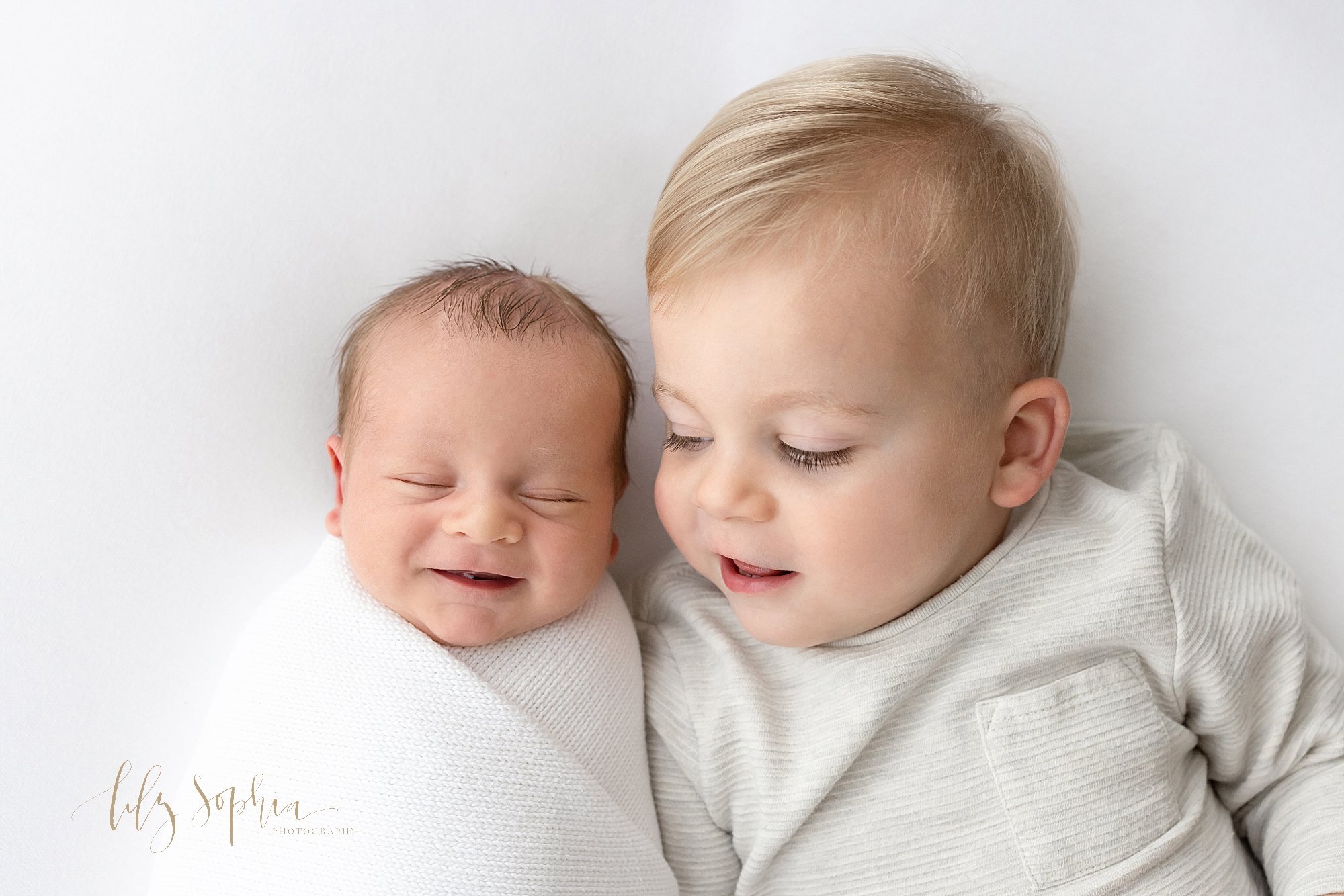  Newborn sibling photo of a toddler boy lying on his back next to his newborn baby brother as his brother smiles in his sleep taken using natural light near Smyrna in Atlanta in a photography studio. 