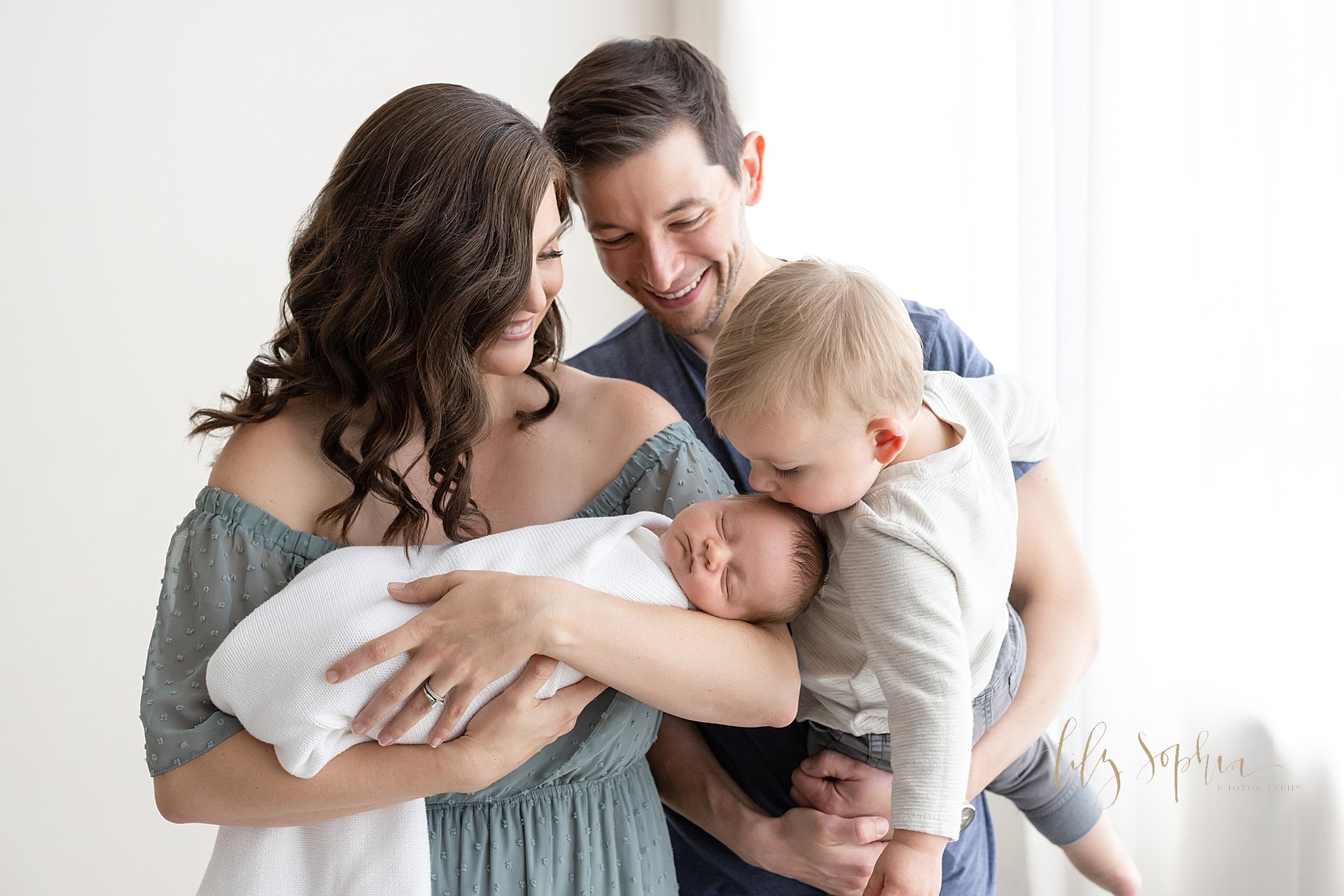  Family newborn portrait of a mother cradling her newborn baby boy in her arms as her husband stands behind and to the left of his wife as he holds their toddler son in his arms and their son kisses his brother on the head taken next to a window stre