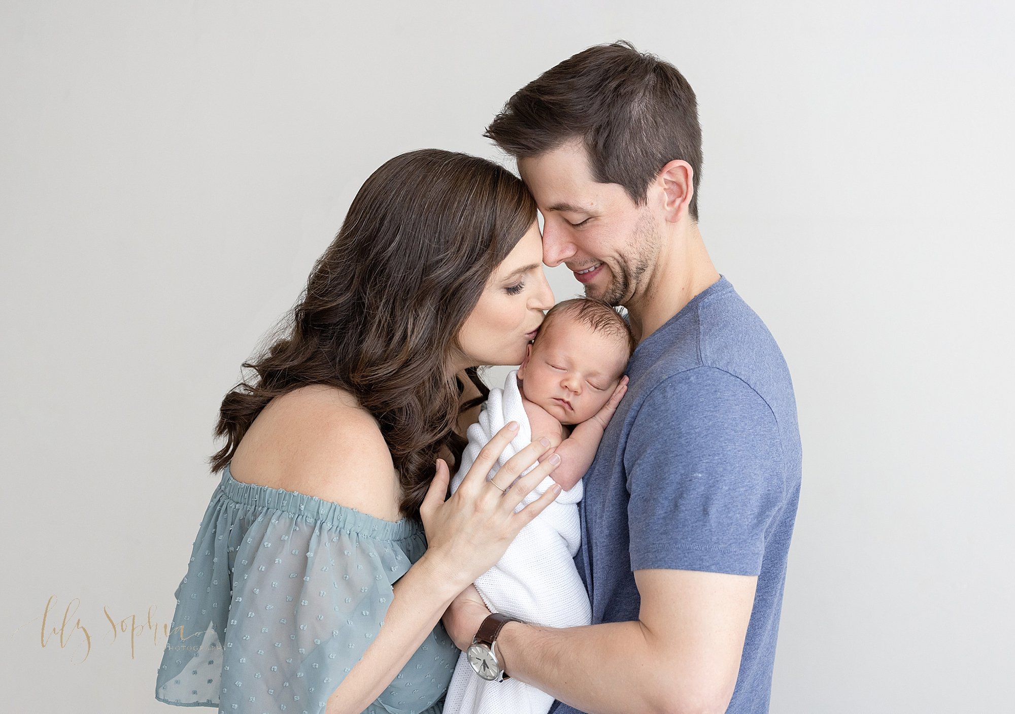  Newborn picture of a father holding his newborn baby son on his chest as his wife faces him and places her hands on her son’s shoulders while she kisses the side of her son’s head as the couple stand in a photography studio near Brookhaven in Atlant