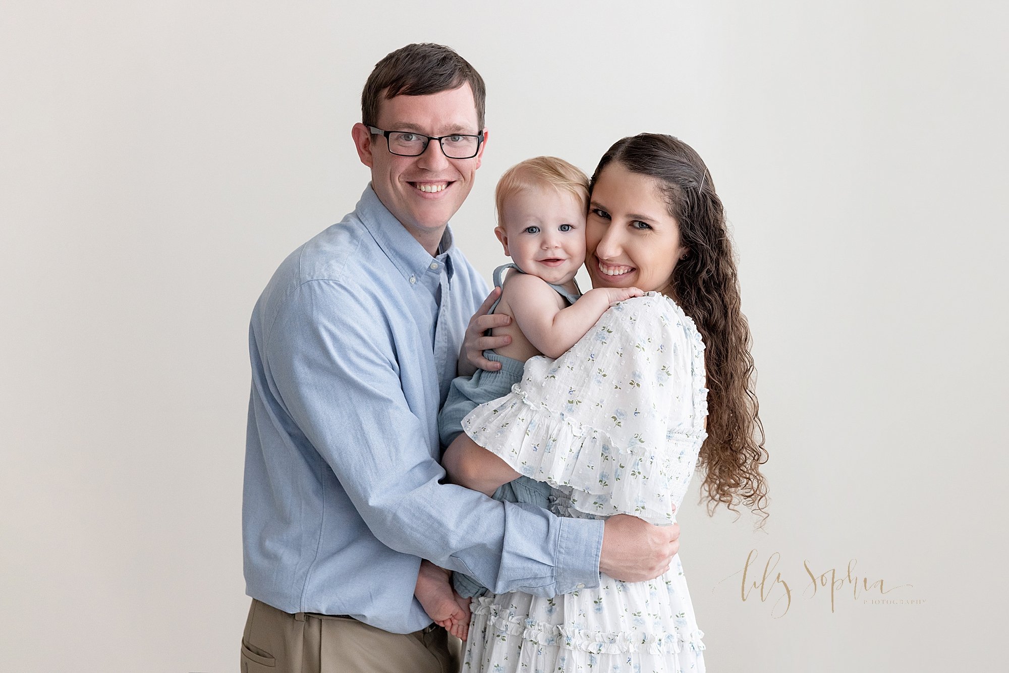  First birthday family portrait of a mother holding her one year old son in her arms with their faces touching one another as her husband faces them and places his hands around her waist taken in a natural light photography studio near Smyrna in Atla