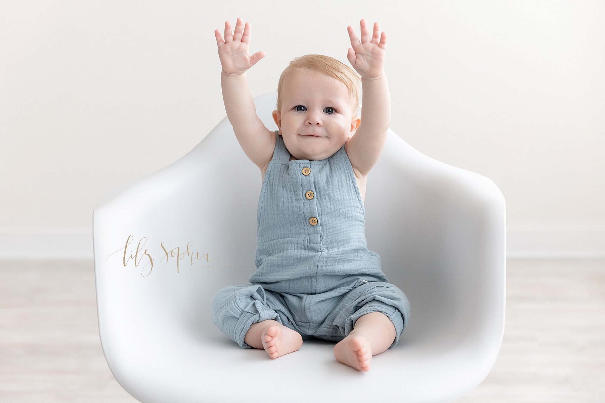  First birthday photo session of a one year old little boy as he gives the touchdown symbol while sitting in a white molded chair taken in a studio using natural light near Vinings in Atlanta. 