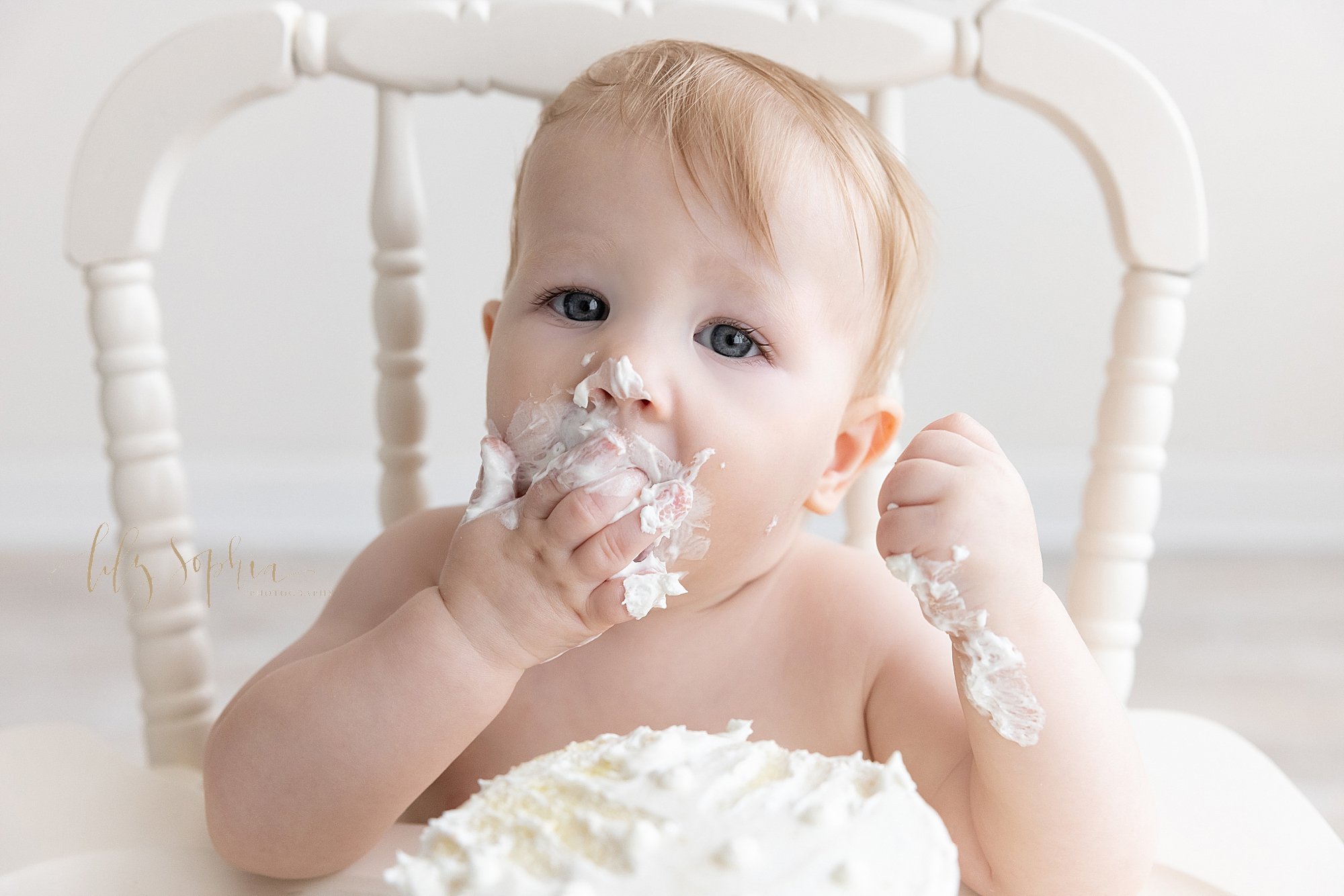 First birthday smash cake photo of a one year old boy as he shoves his cake into his mouth while sitting in an antique high chair in a photography studio using natural light located near Brookhaven in Atlanta, Georgia. 