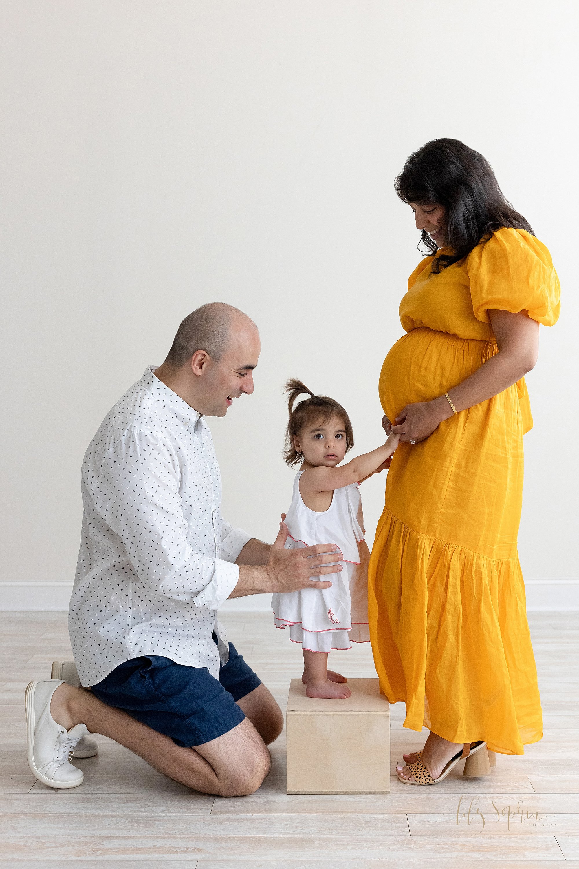  Family maternity photo session in a natural light studio of a pregnant mother standing facing her husband who is kneeling behind his young daughter who is holding the hands of her mother as her father supports her with his hands  at her waist while 