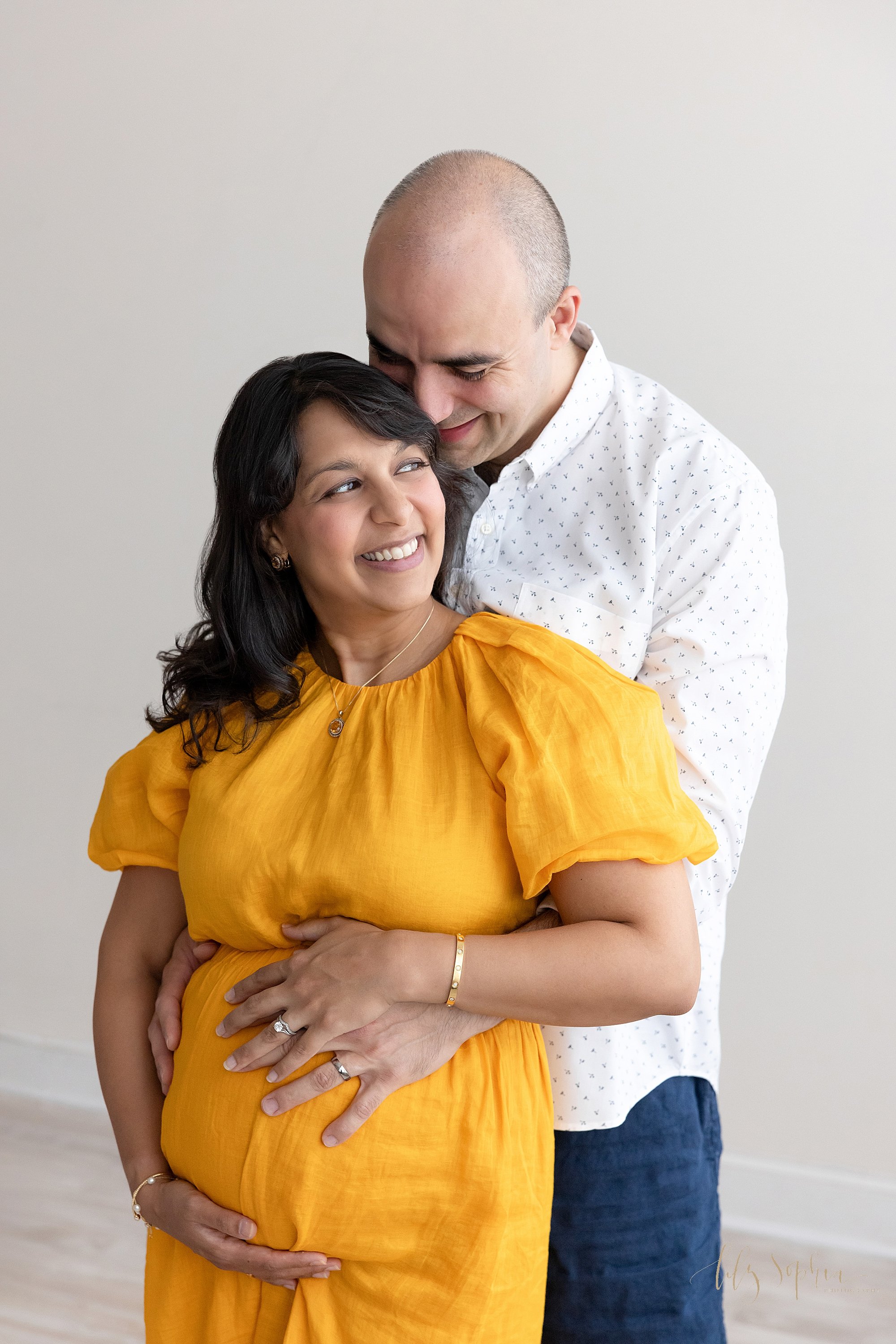  Maternity picture taken in a photography studio with the husband standing behind his pregnant wife as she holds the base of her belly with her right hand and her husband places his left hand on their baby in utero as his wife puts her hand on top of