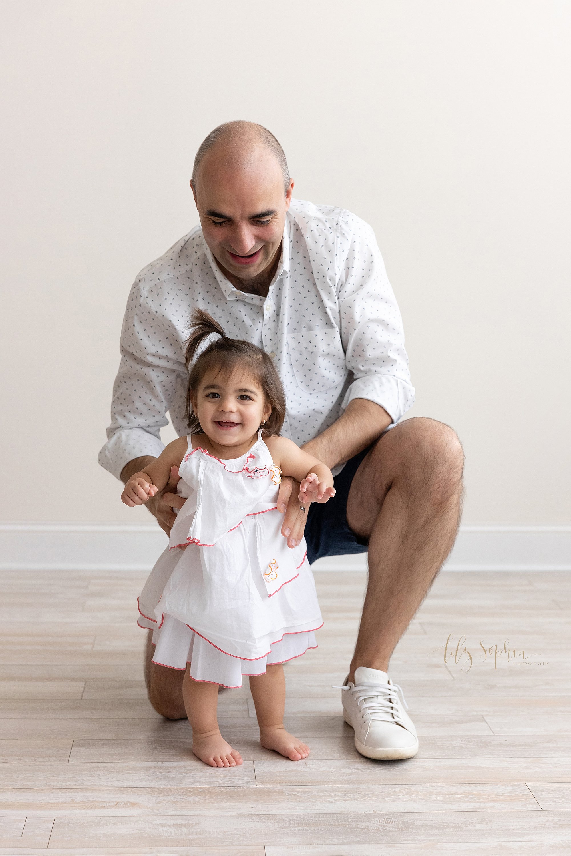  Family photo of a father squatting behind his toddler daughter as he supports her to walk in a photography studio that uses natural light near Poncey Highlands in Atlanta. 