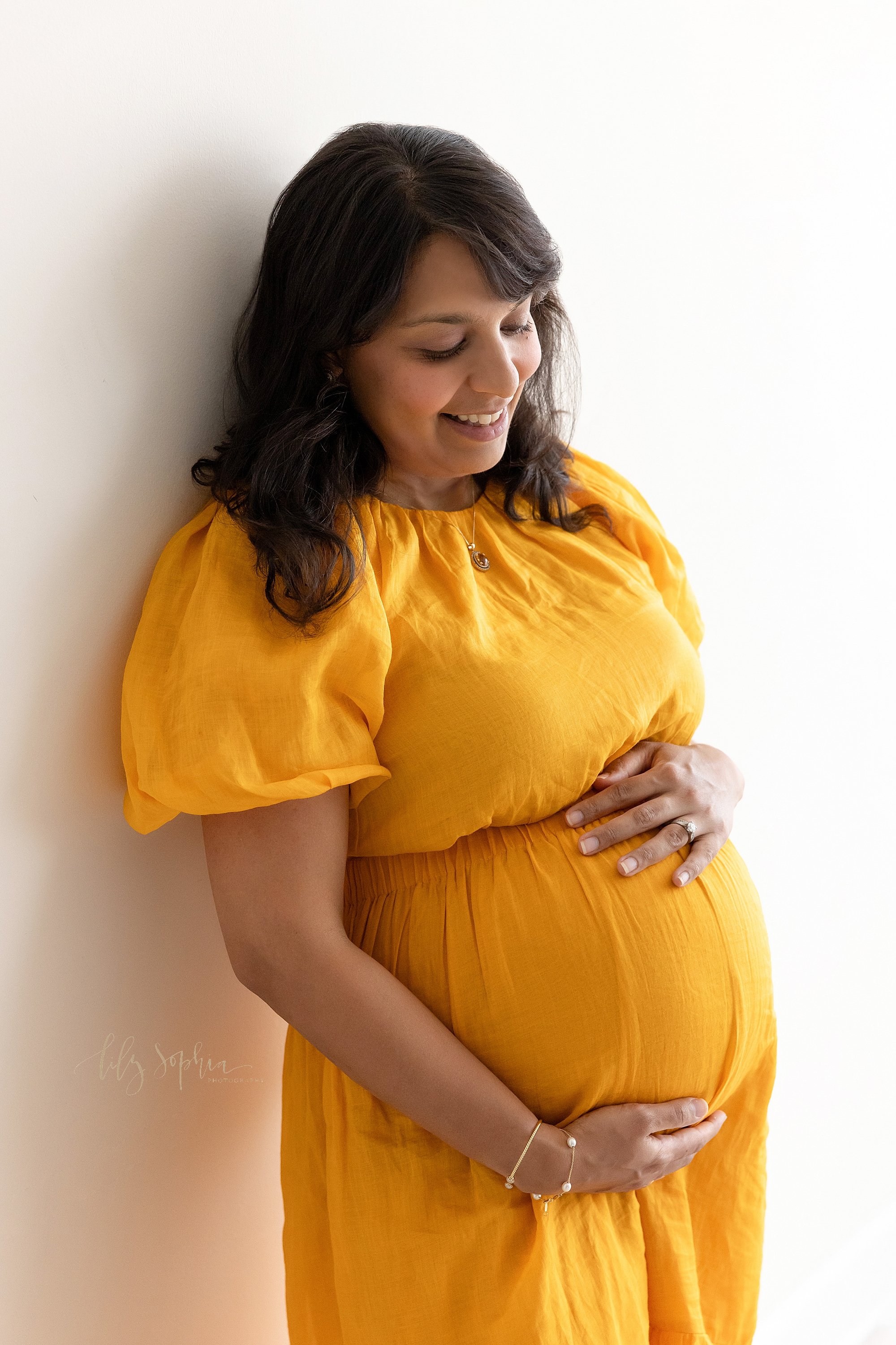  Maternity portrait of a pregnant mother standing with her back against the wall of a photography studio as she frames her belly with her hands while standing next to a window streaming natural light as she contemplates the upcoming birth of her chil