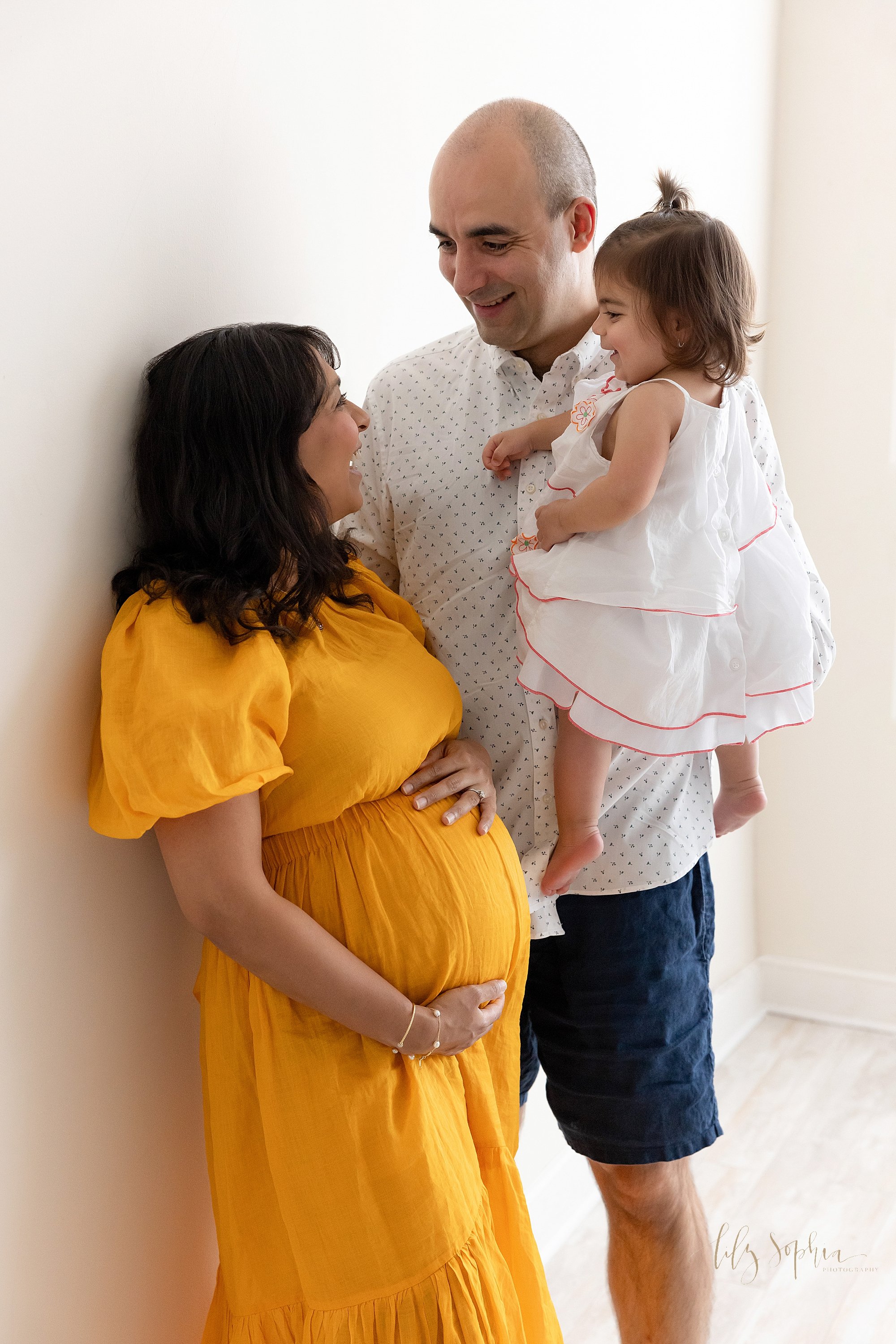  Family maternity photo shoot with an expectant mother framing her belly with her hands as she turns her head over her left shoulder to talk with her husband who is standing with his right shoulder to the wall of the studio and holding his toddler da