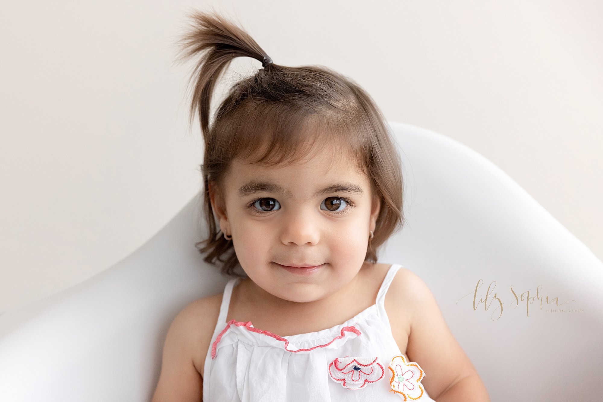  Family photo of a toddler girl wearing a pebbles ponytail on top her head as she sits in a molded white chair in a natural light photography studio near Kirkwood in Atlanta. 