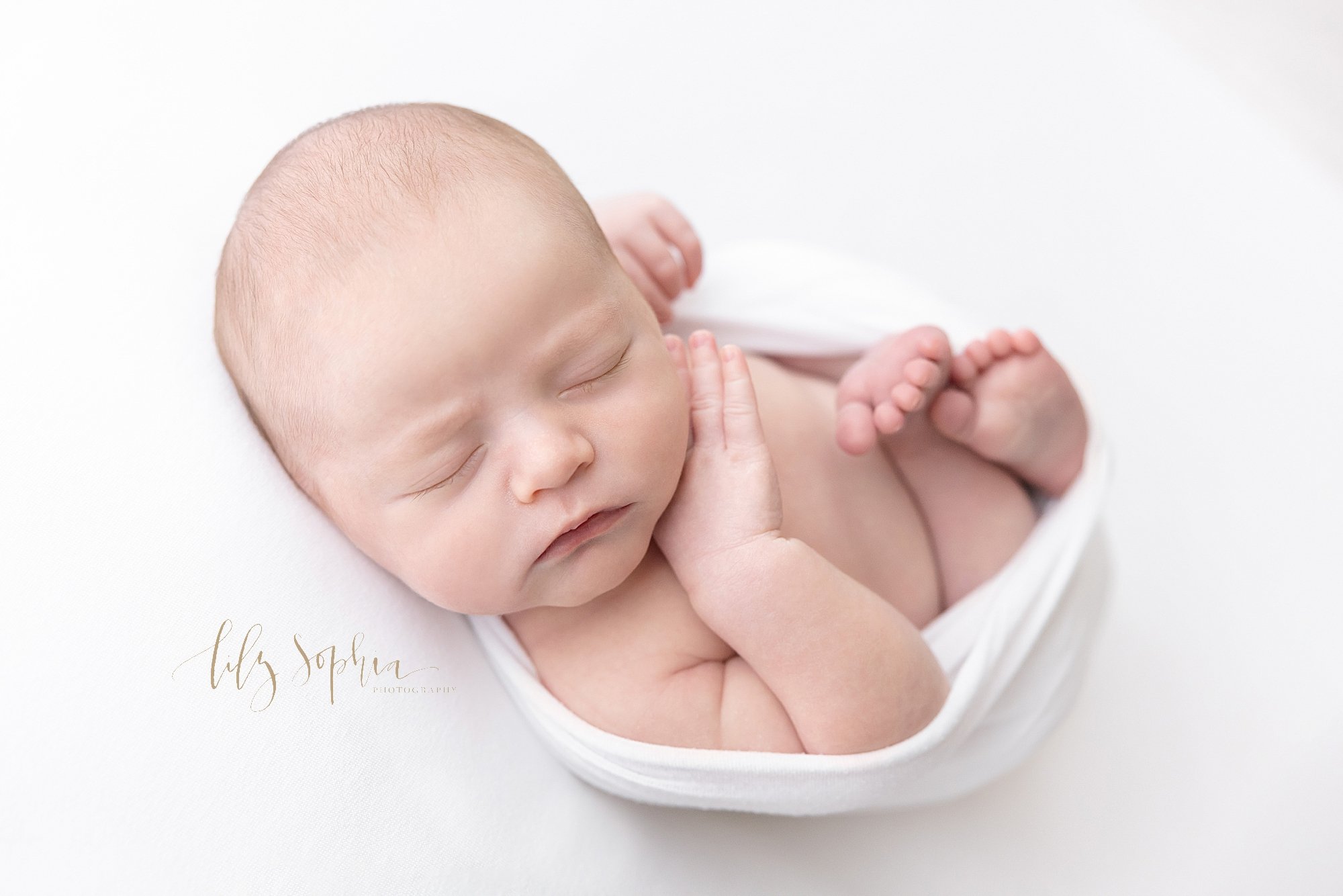  A bundle of newborn joy is captured in this newborn picture of a baby boy cradled in a stretchy swaddle with his right hand under his chin taken using natural light near Sandy Springs in Atlanta in a photography studio. 