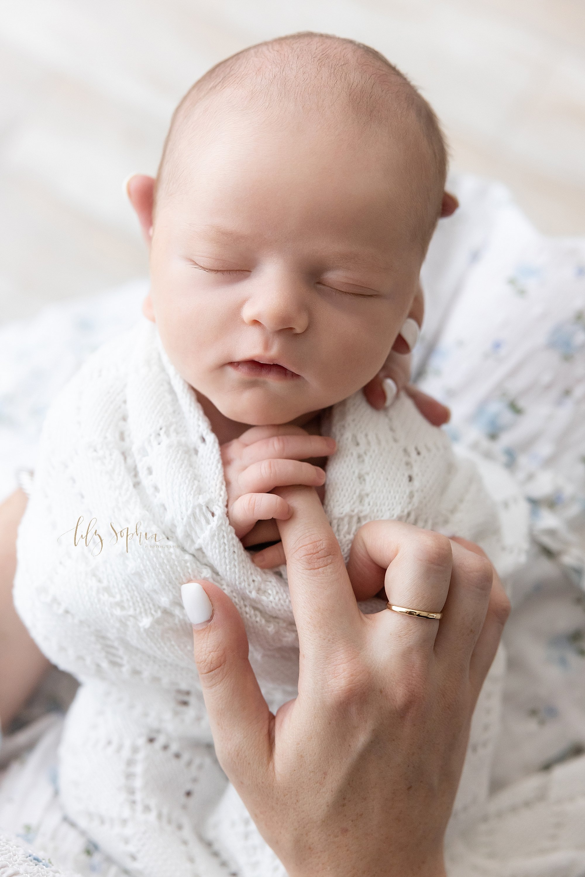  Close-up newborn picture of a newborn baby boy wrapped in a soft white knitted blanket as his head is head in the left hand of his mother and her son holds her right index finger taken in a natural light photography studio in Ponce City Market in At