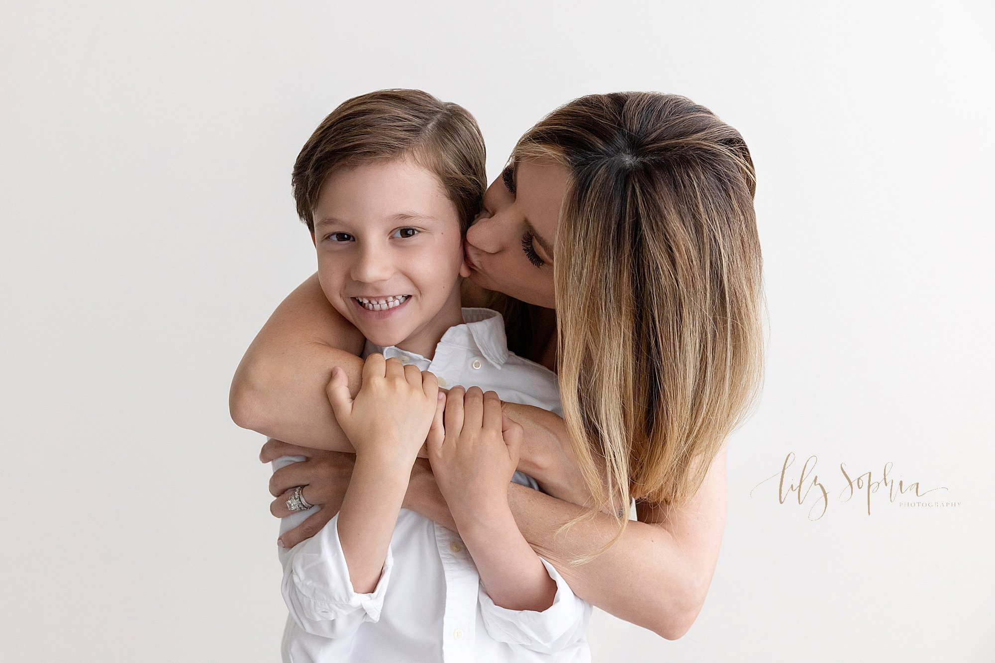  Family photo of young boy standing in a studio while his mother wraps his arms around his chest and he hangs onto her right arm while she kisses him taken in natural light near Roswell in Atlanta, Georgia. 