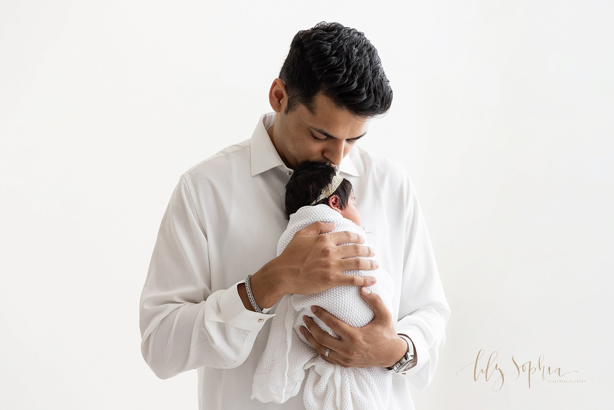  Newborn picture of a newborn baby girl being held by her father against his chest as he kisses the crown of her head while standing next to a window streaming natural light in a studio near Old Fourth Ward in Atlanta. 