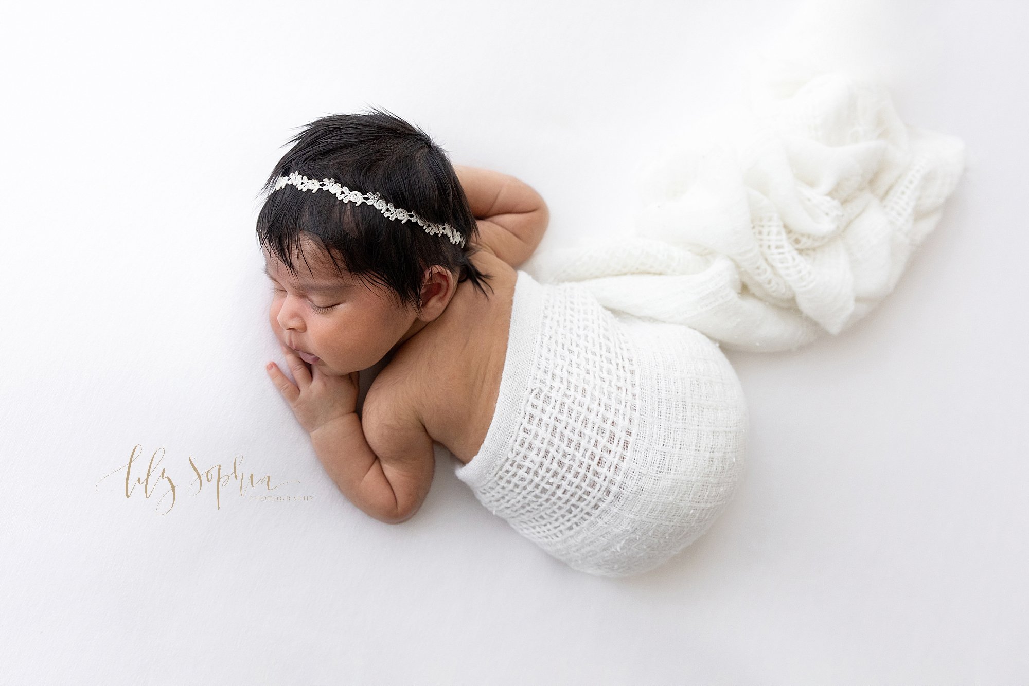  Newborn portrait of a sleeping newborn baby Indian girl as she lies on her stomach with her head turned to her right wearing a delicate headband in her jet black hair and wrapped in a soft white blanket taken near Morningside in Atlanta in a studio 