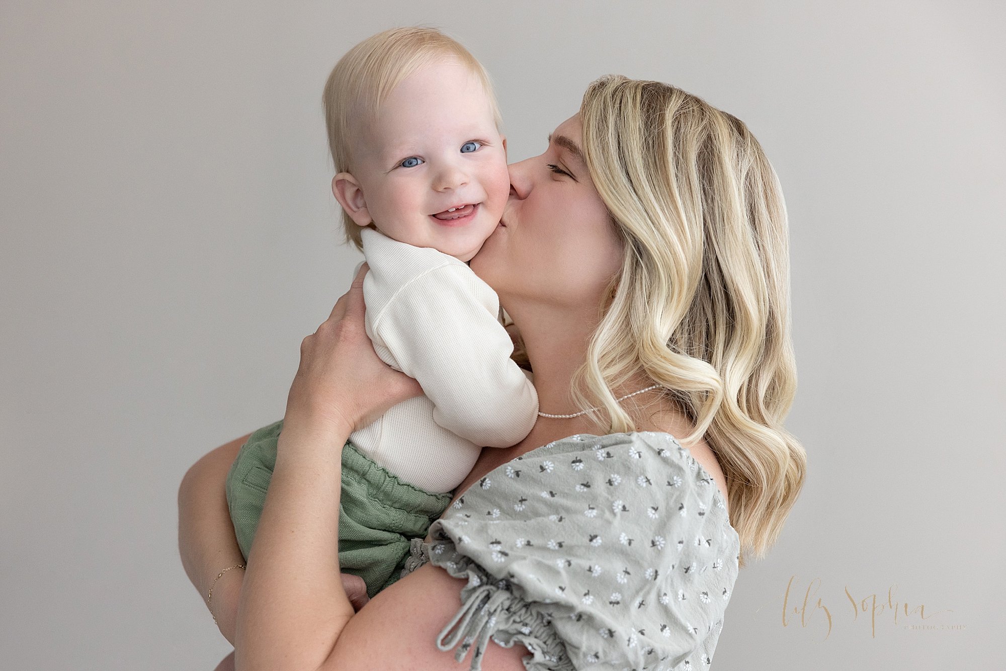  First birthday family photo of a mother holding her one year old son and kissing him on his neck to make him smile so he can show his tiny teeth taken in a photography studio that uses natural light near Oakhurst in Atlanta. 