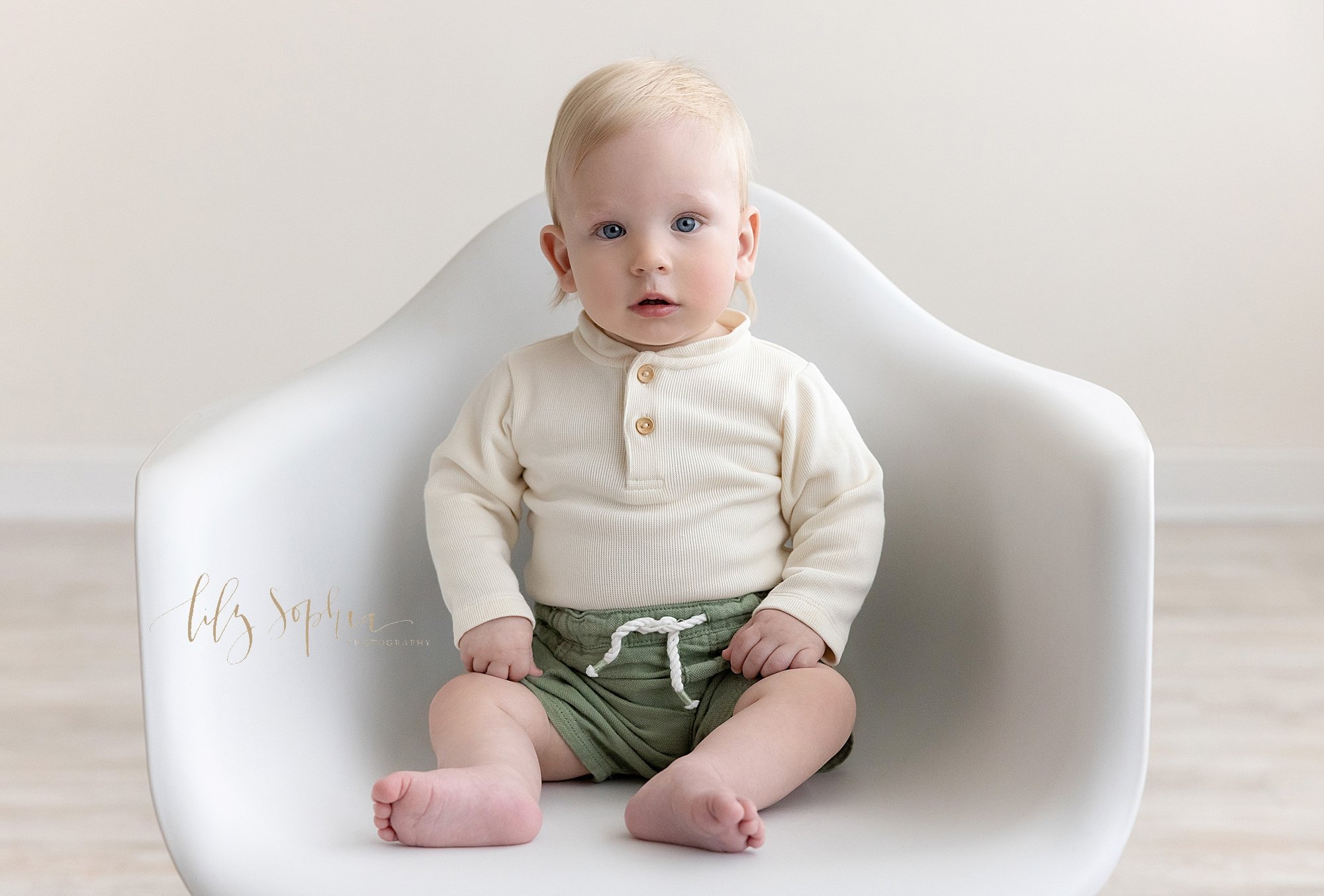  First birthday picture of a one year old boy sitting in a white molded chair taken next to a window streaming natural light in a photography studio near Cumming in Atlanta, Georgia. 