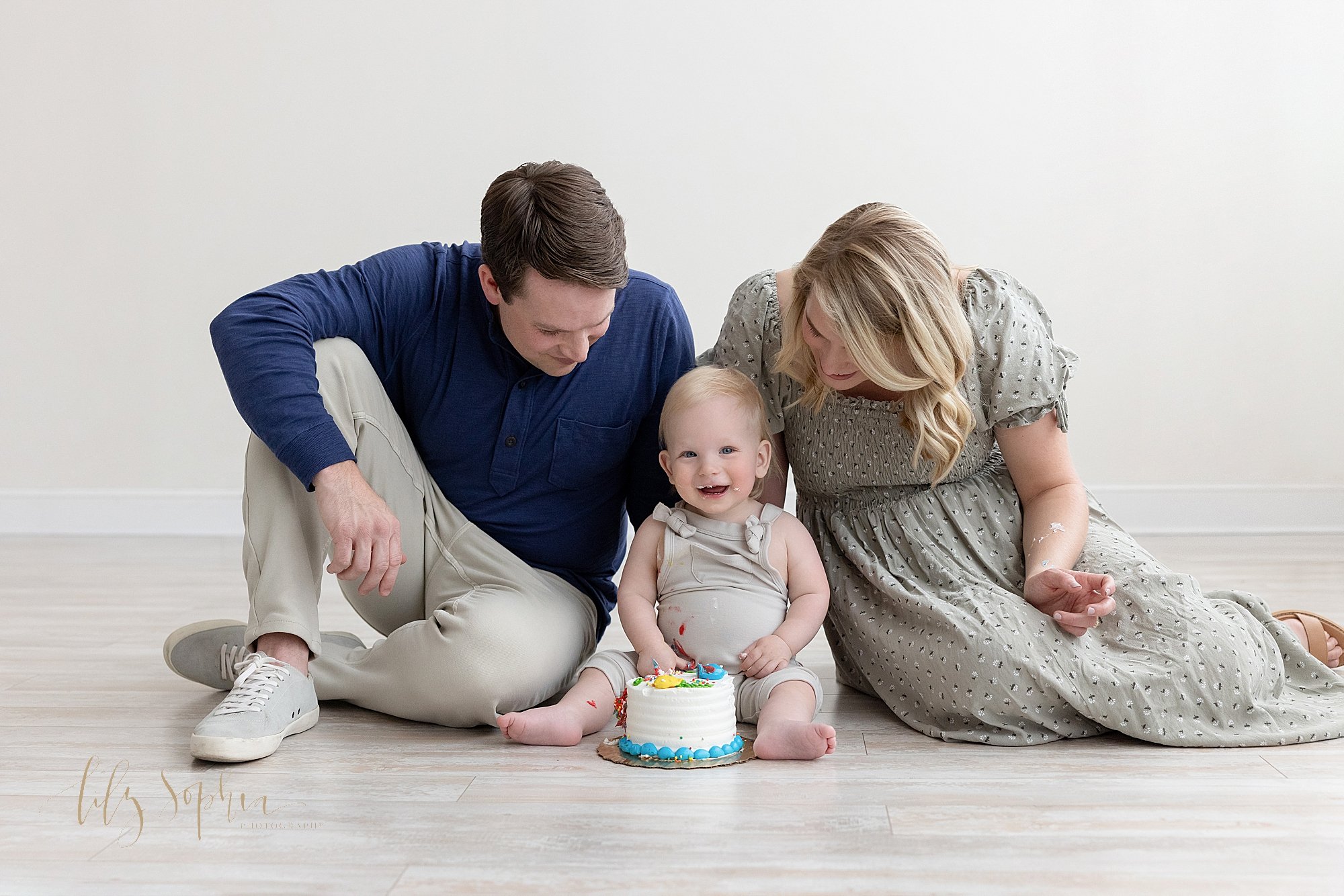  Family first birthday smash cake photo of a mother and father sitting on the floor of a photography studio with their one year old son between them as he straddles his smash cake taken using natural light near Ansley Park in Atlanta. 