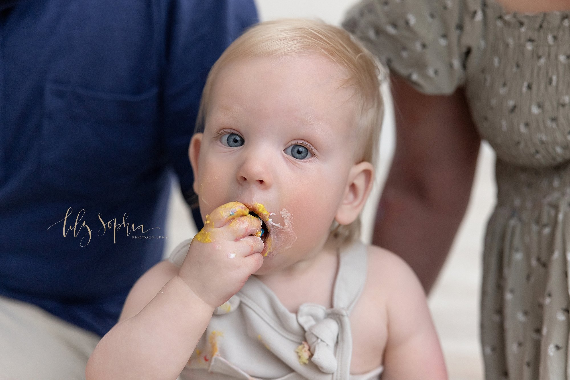  First birthday smash cake photo of a one year old boy sitting on the floor of a natural light studio between his parents as he shoves his cake into his mouth with his right hand taken near Alpharetta in Atlanta. 