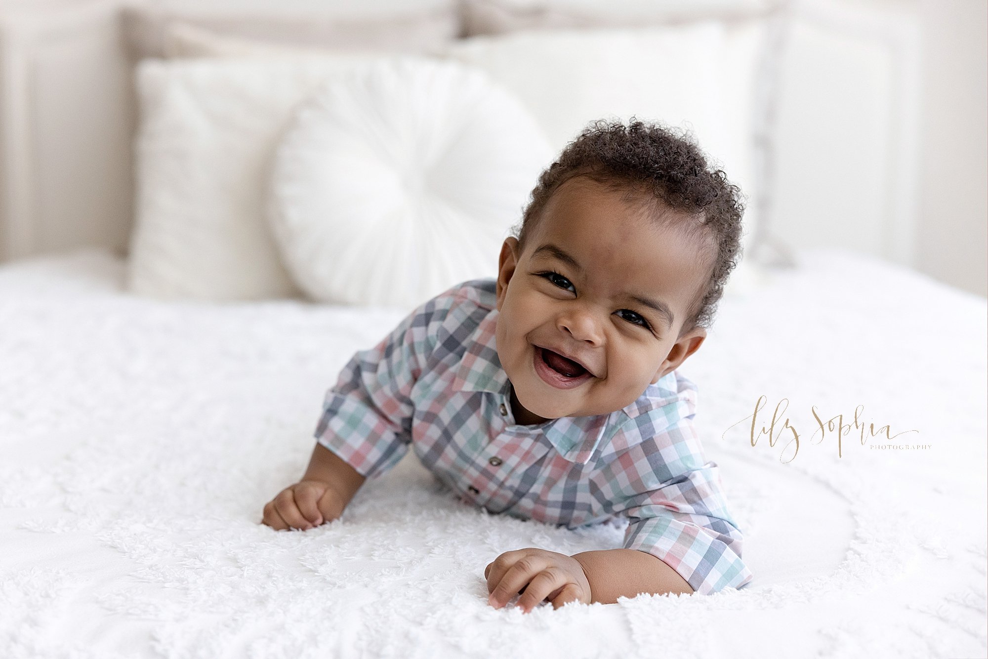  First birthday photo session of an African-American one year old boy as he laughs while crawling across a bed in a studio near Virginia Highlands in Atlanta that uses natural light. 