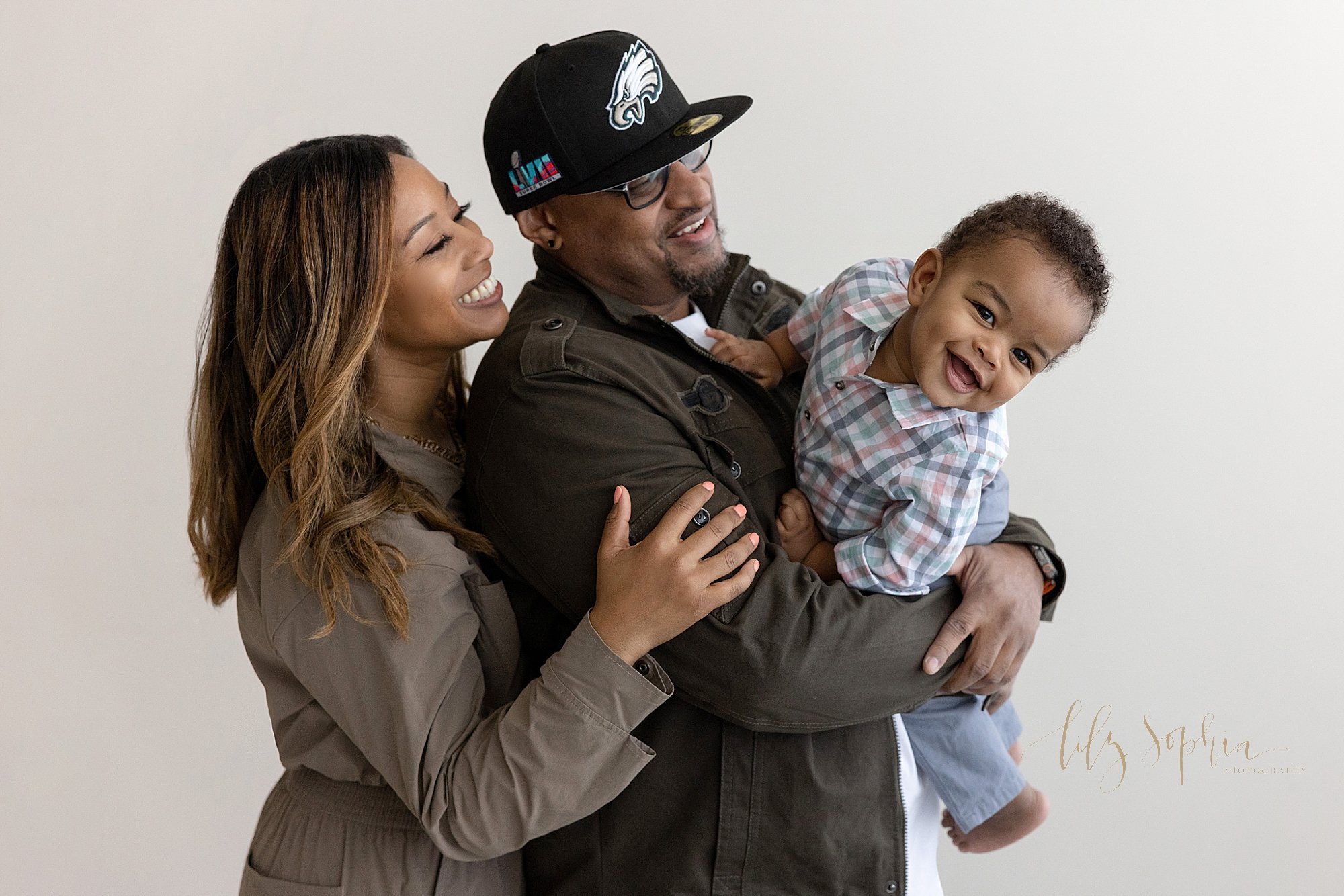  Family first birthday portrait of an African-American father holding his one year old in his arms as he tries to squirm to get down and his mother stands behind his father looking on taken next to a window streaming natural light in a photography st