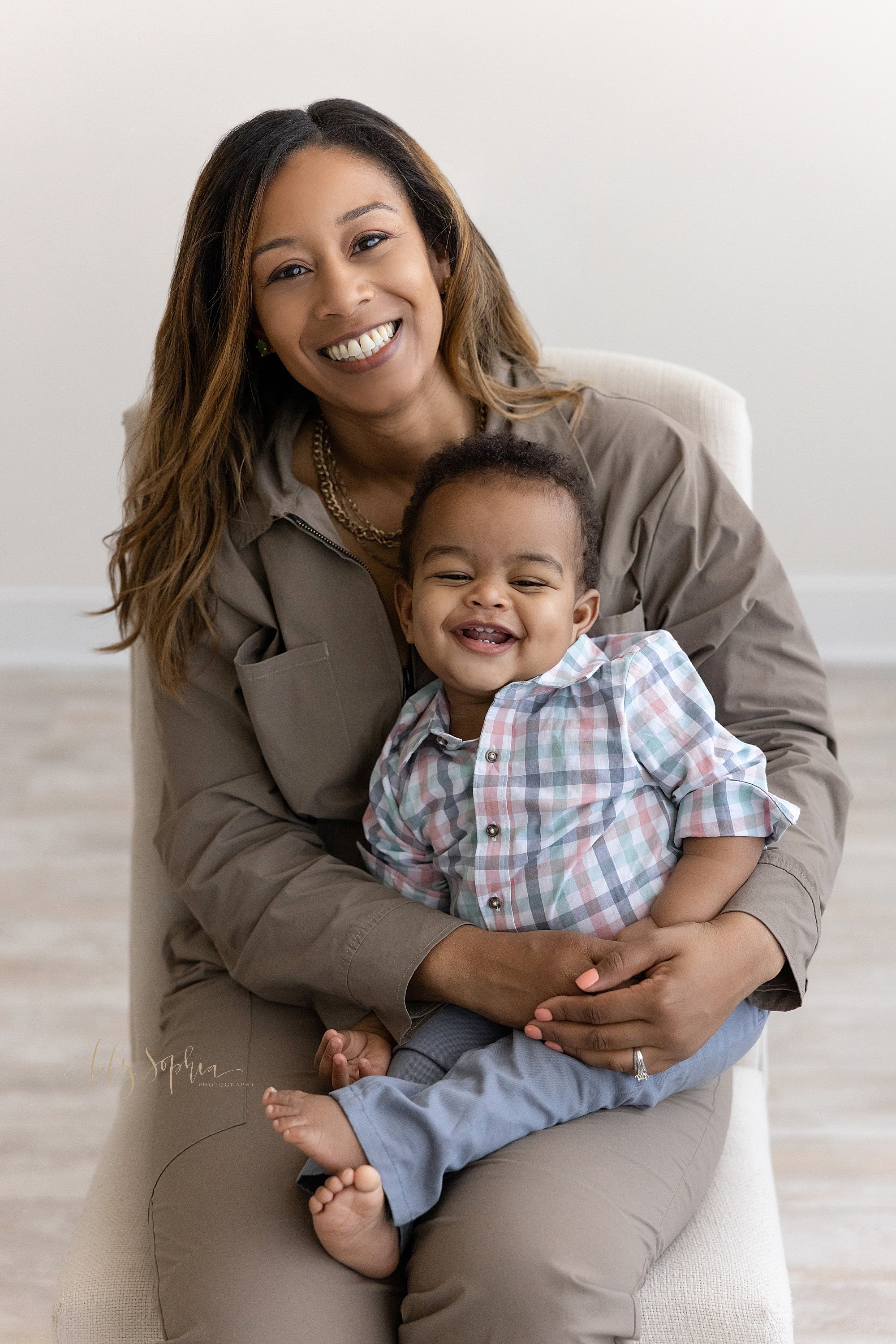  First birthday family photo shoot with a one year old African-American boy giggling to show his tiny teeth as he sits on his mother’s lap next to a window streaming natural light in a photography studio near Poncey Highlands in Atlanta, Georgia. 