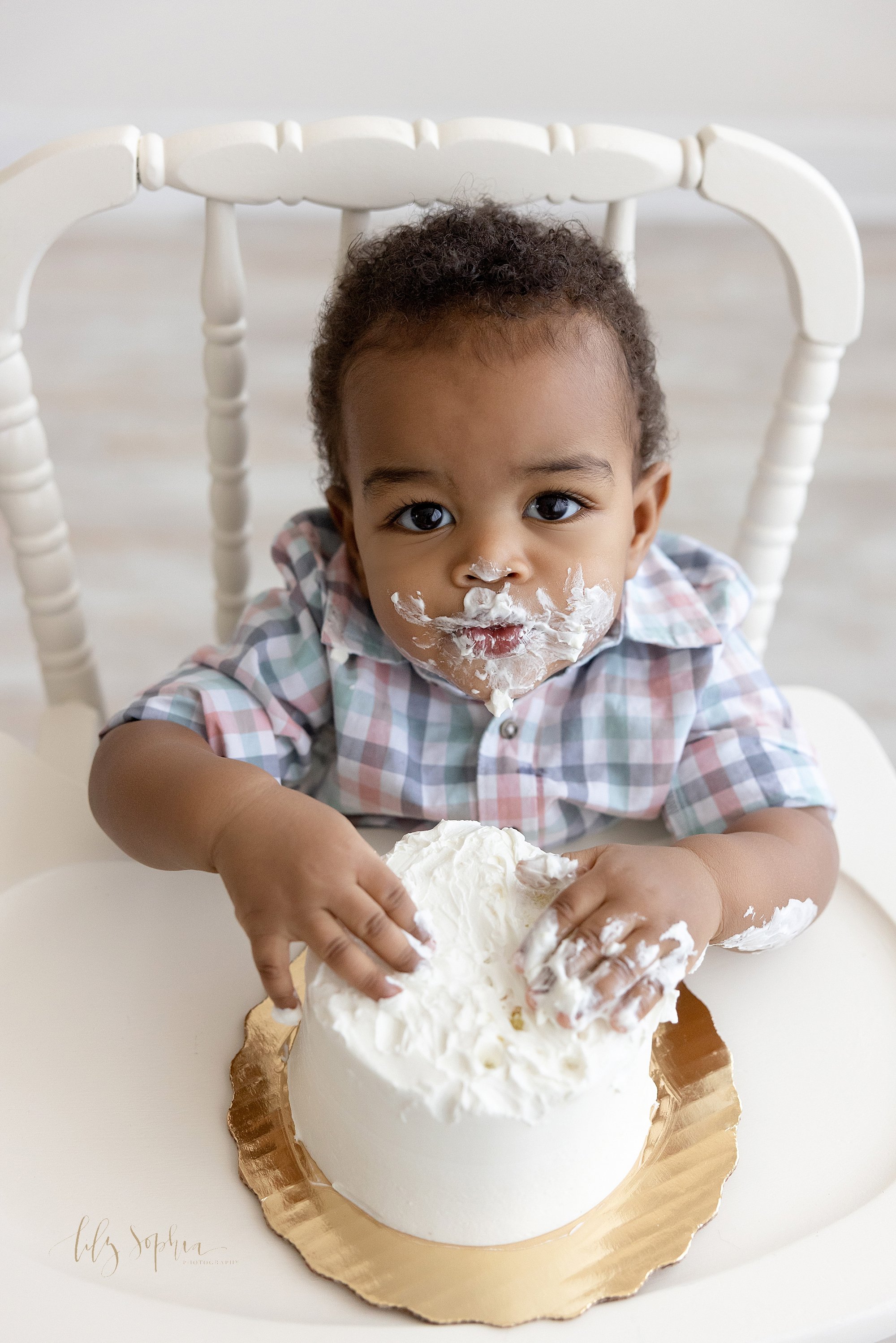  Smash cake first birthday picture of an African-American one year old boy sitting in an antique highchair with icing covering his face and his hands digging into his cake taken near Brookhaven in Atlanta in a natural light photography studio. 