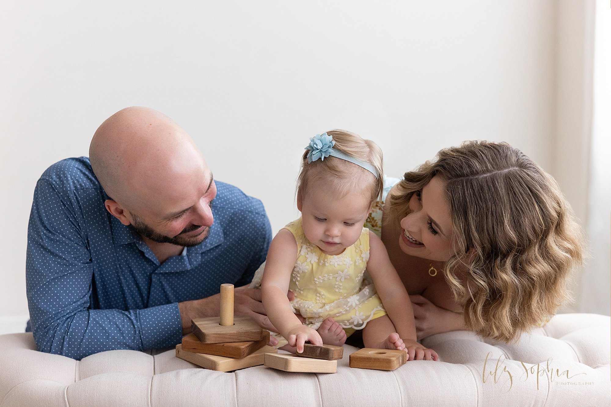  Family photo of a one year old girl sitting on top of a tufted bench playing with a wooden stacking toy as her parents watch while they kneel behind the bench taken in a natural light studio near Ansley Park in Atlanta, Georgia. 