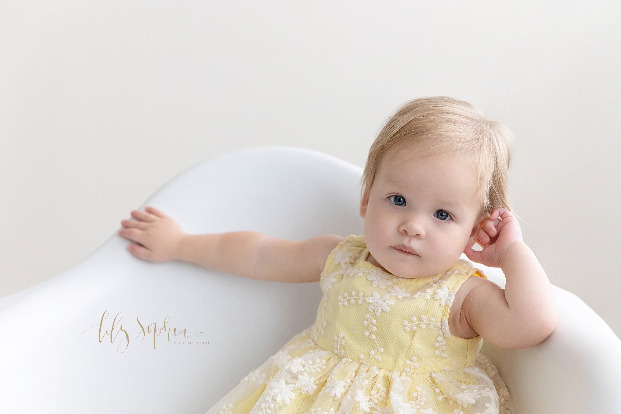  First birthday photo session of a one year old girl as she sits in a white molded chair with her left hand propping up her head and her right arm extended to hold the back of the chair taken next to a window streaming natural light in a studio near 