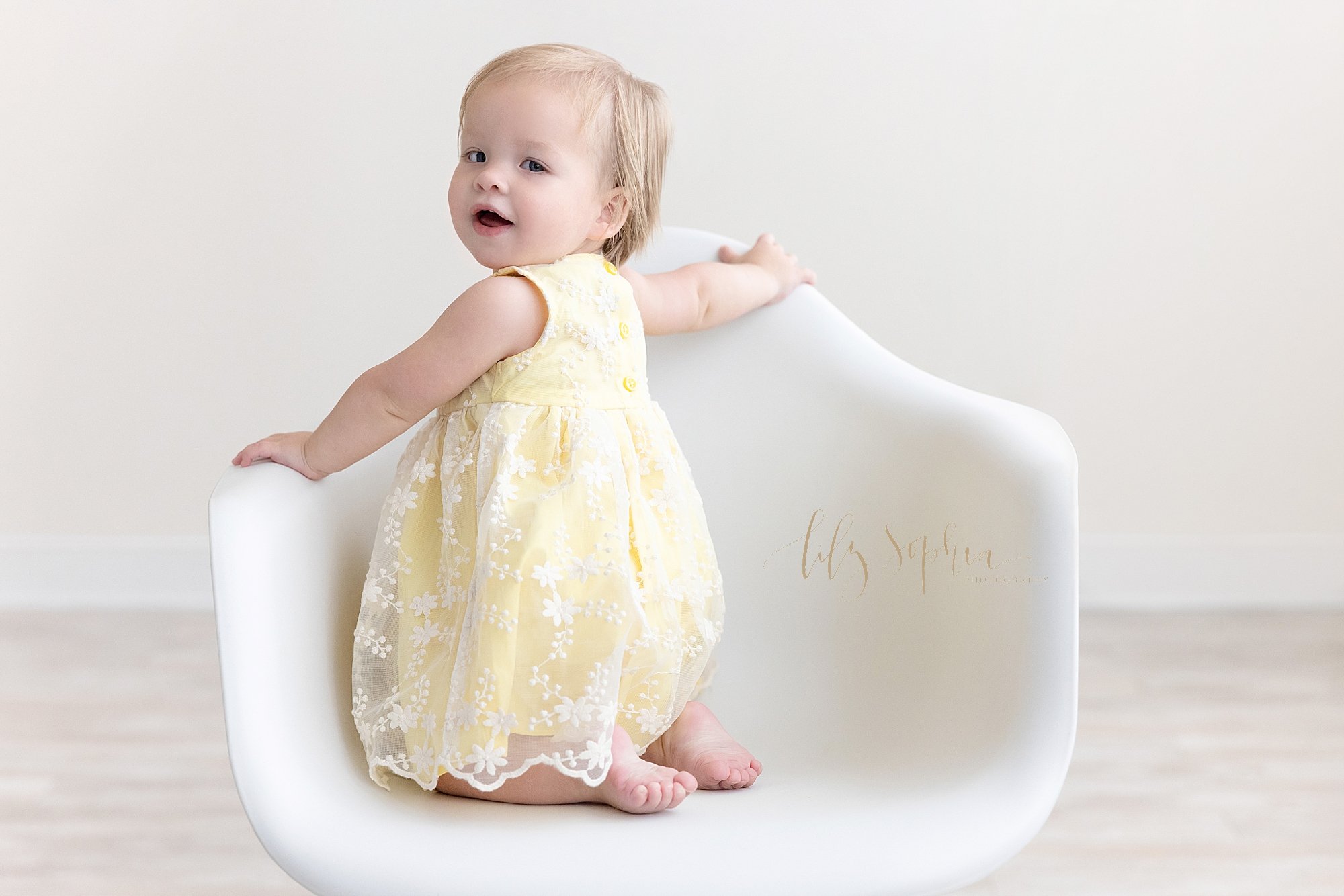  First birthday photo of a one year old little girl who is kneeling in a white molded chair as she looks over her left shoulder taken in a studio near Morningside in Atlanta that uses natural light. 