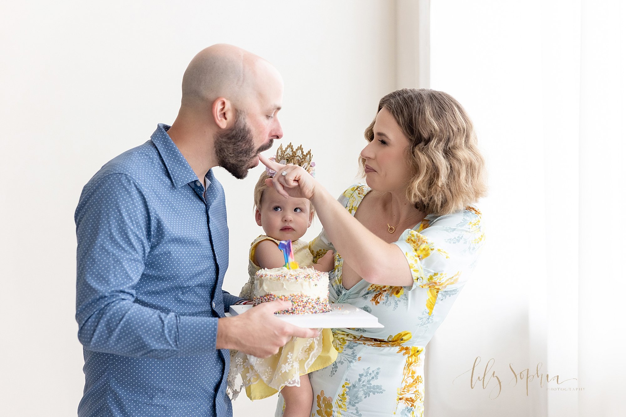  Family first birthday photo session with a father holding a smash cake in front of his one year old daughter who is being held by her mother as mom gives dad a lick of the icing as all three are next to a window streaming natural light into a photog