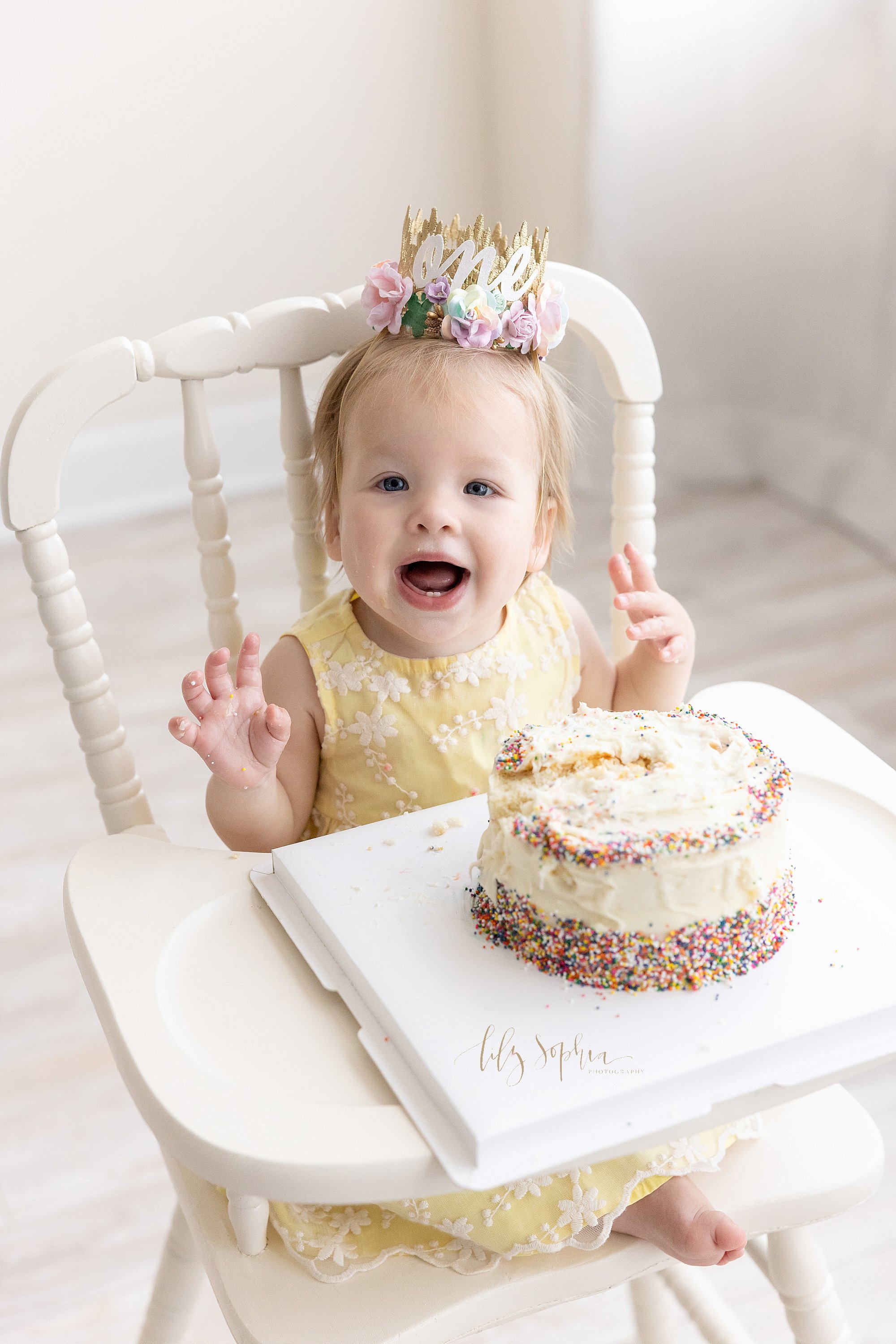  First birthday smash cake photo of a one year old girl wearing a crown on her head excited to be eating her birthday cake while seated in an antique highchair taken near Brookhaven in Atlanta in a natural light photography studio. 