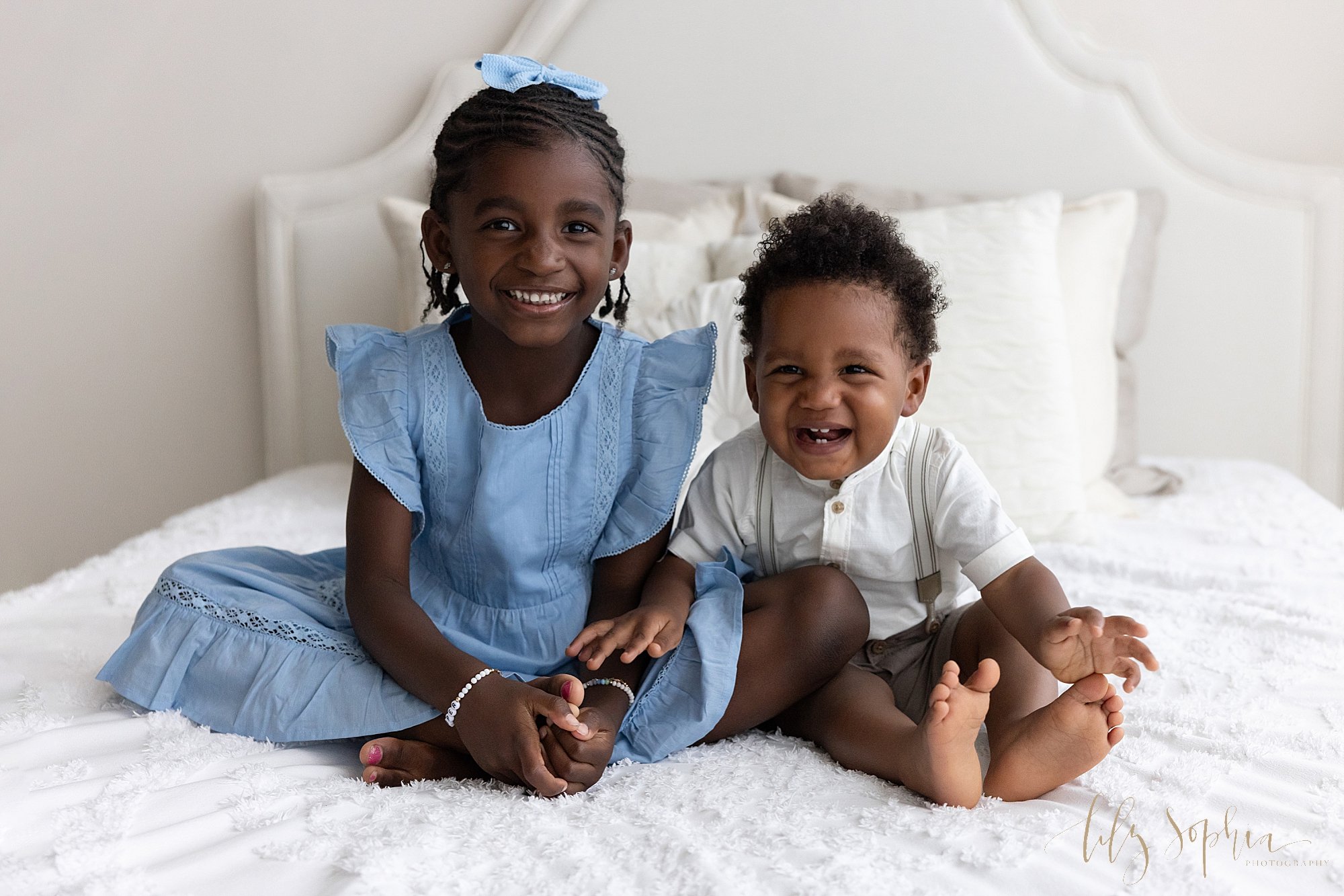  Family photo session of African-American siblings, a young girl and a baby boy as they sit next to one another atop a bed next to a window streaming natural light in a photography studio near Sandy Springs in Atlanta. 