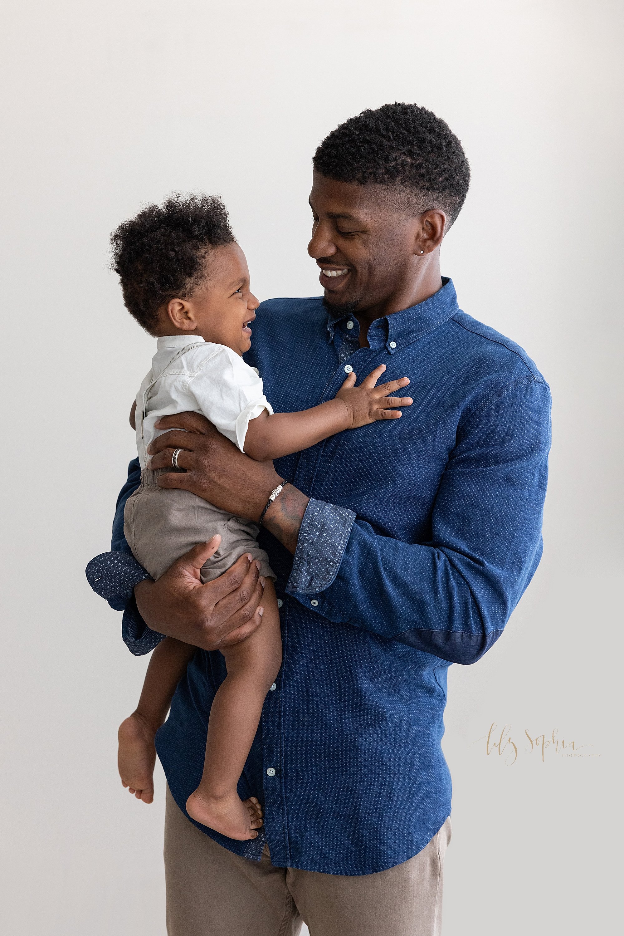  Family father and son portrait of an African-American father holding his toddler son in his arms as the two of them talk and laugh with one another taken in a natural light studio near Virginia Highlands in Atlanta. 