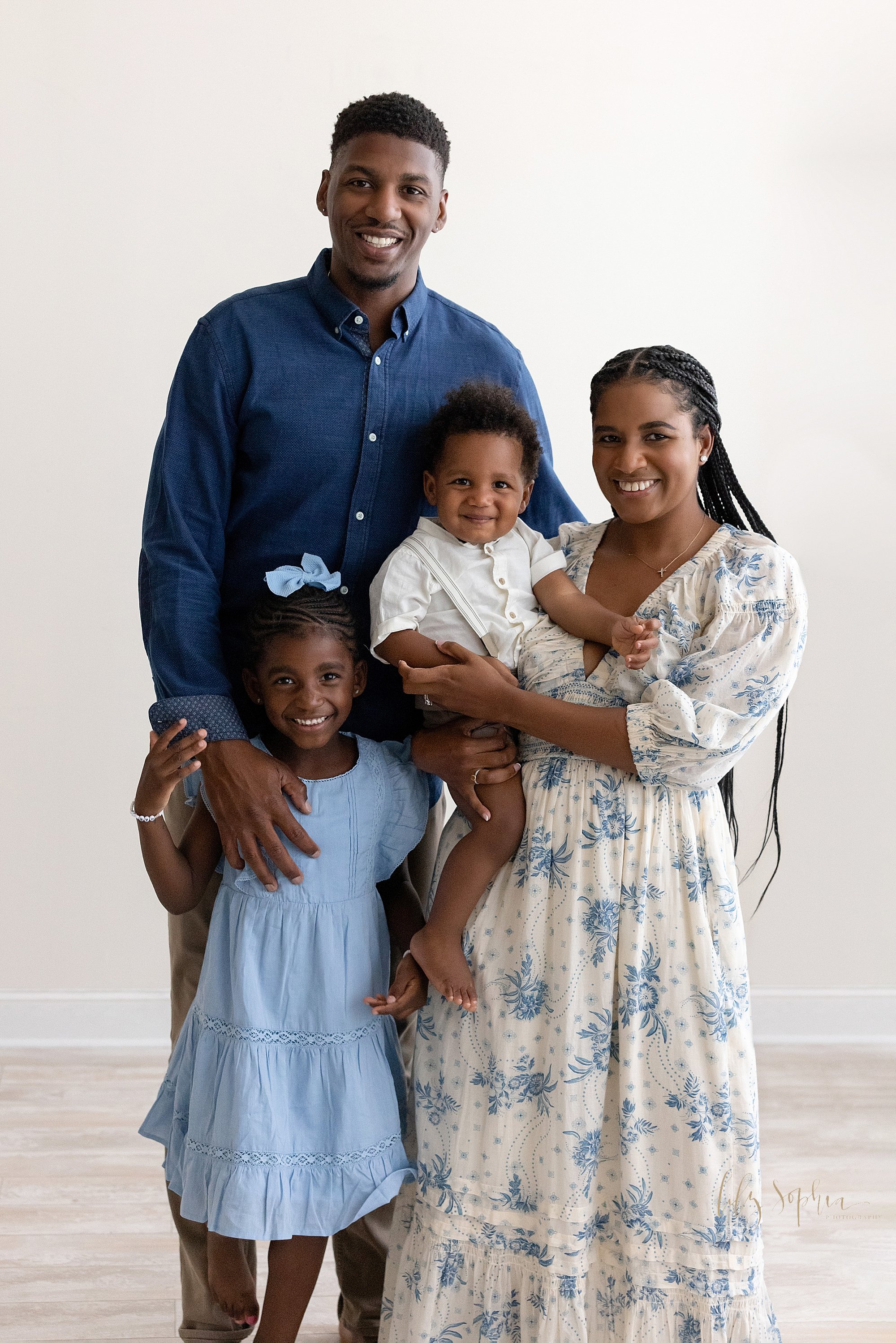  Family portrait of an African-American father standing with his daughter in front of him on his right side and his wife holding their toddler son on his left side next to a window streaming natural light into a photography studio in Ponce City Marke