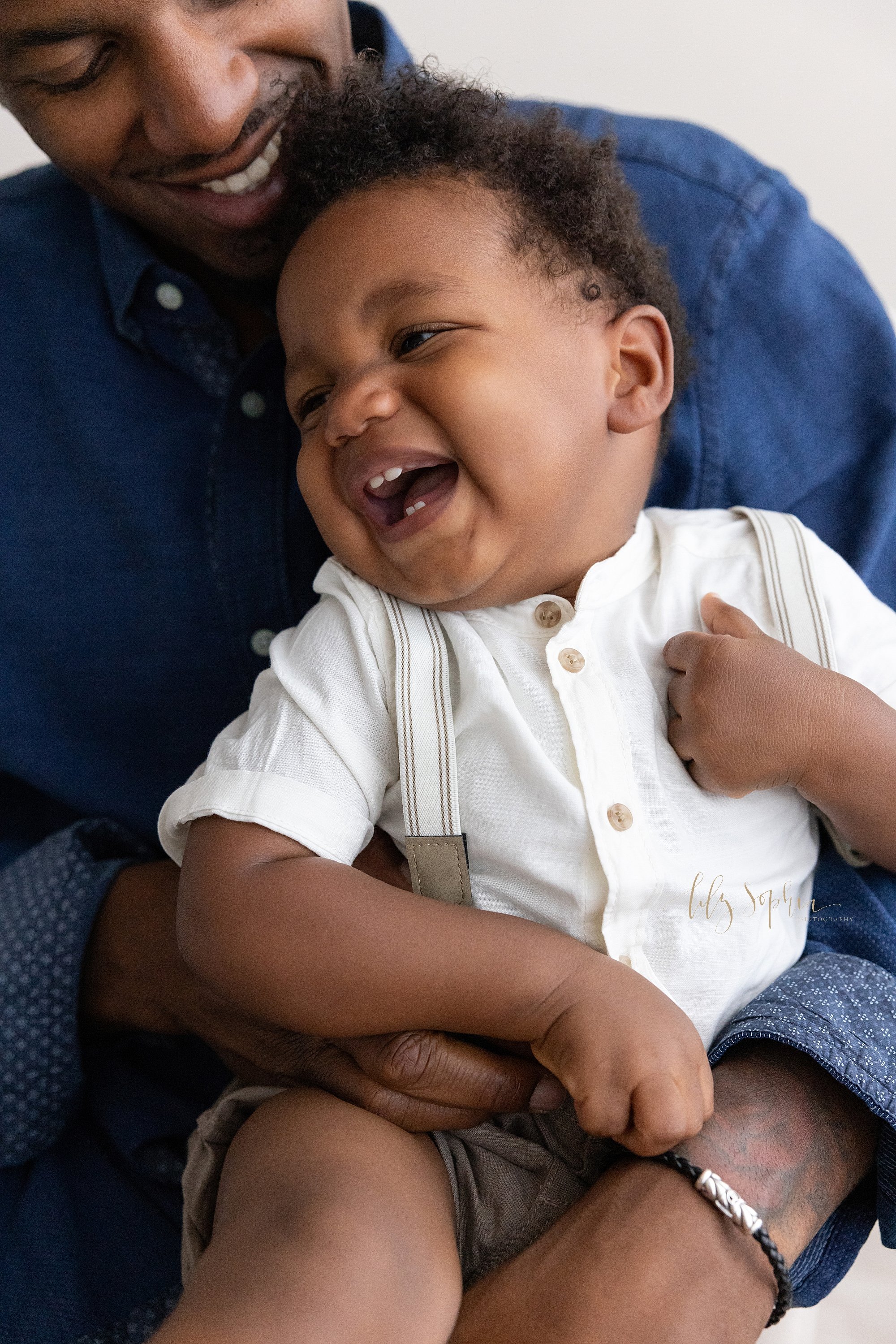  Fun family photo of an African-American father laughing with his toddler son as he holds him on his lap next to a window streaming natural light in a photography studio near Old Fourth Ward in Atlanta. 