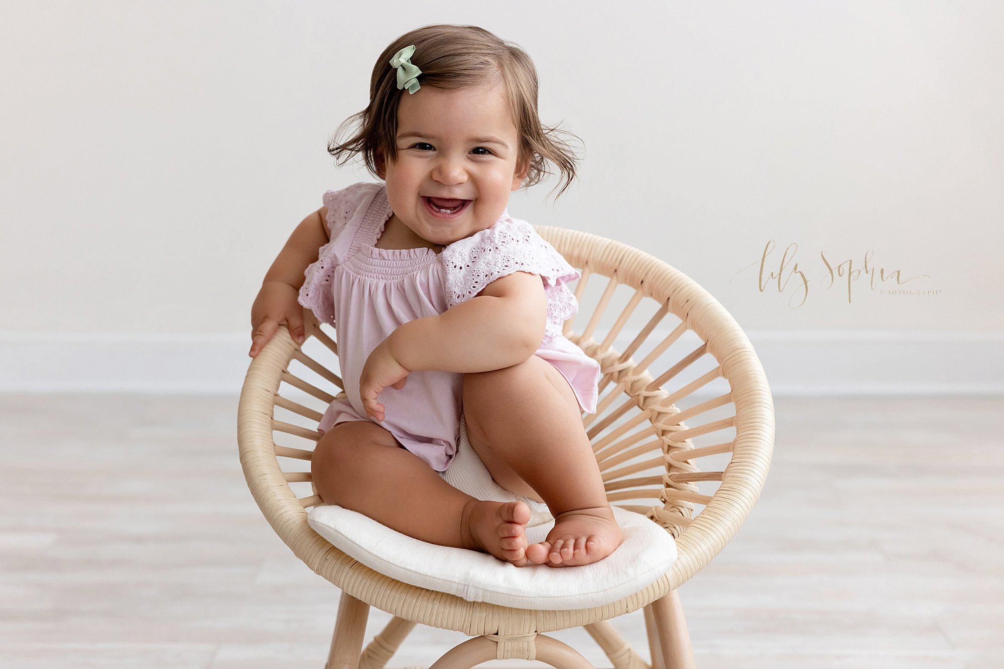 First birthday portrait of a spunky one year old little girl as she sits in a bamboo chair with her left elbow on her left knee and smiles to show her tiny bottom teeth taken near Oakhurst in Atlanta in a natural light studio. 