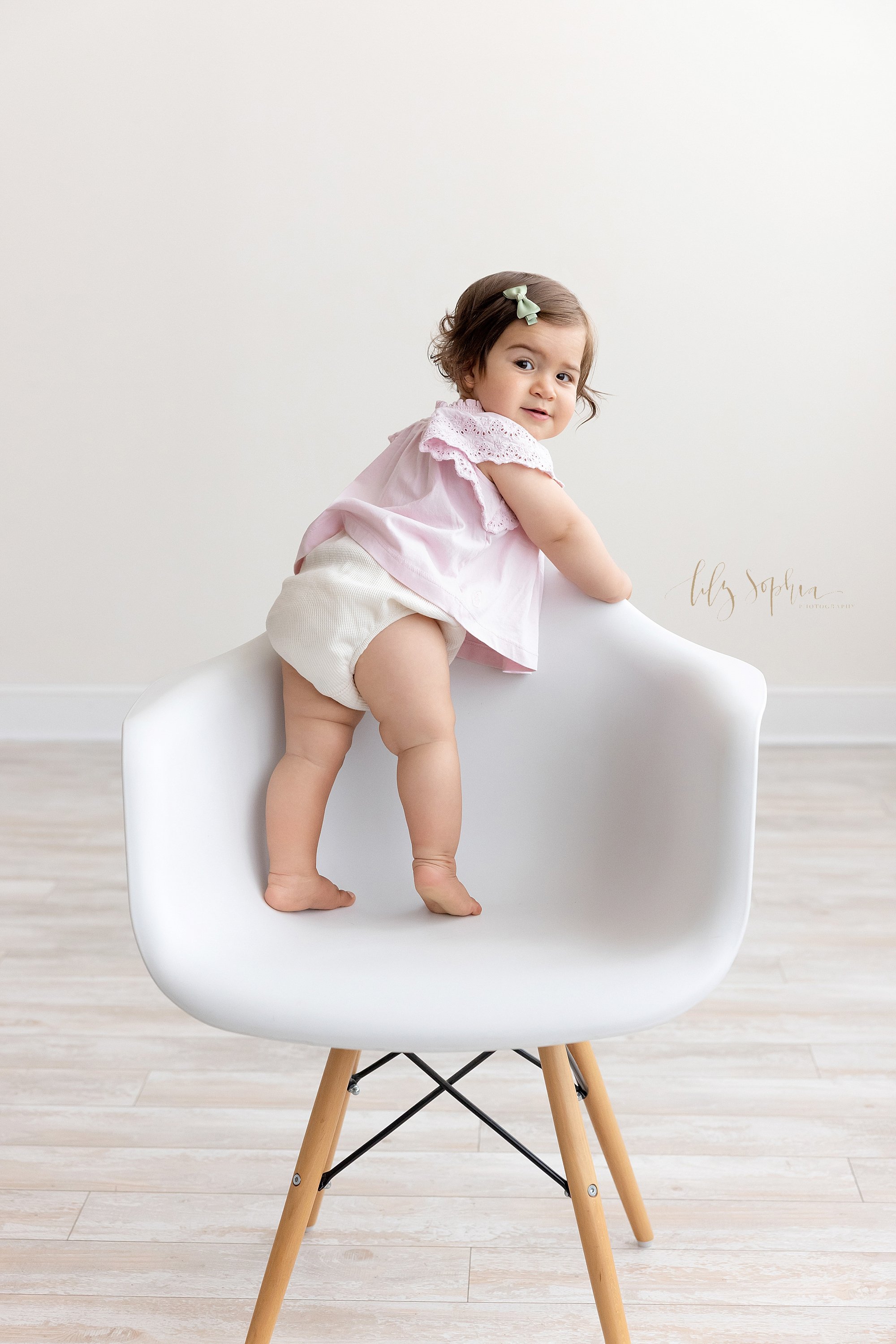  First birthday photo of an adventurous one year old baby girl as she stands up in a white molded chair while holding onto the back of the chair and looks over her right shoulder taken near Virginia Highlands in Atlanta in a photography studio that u