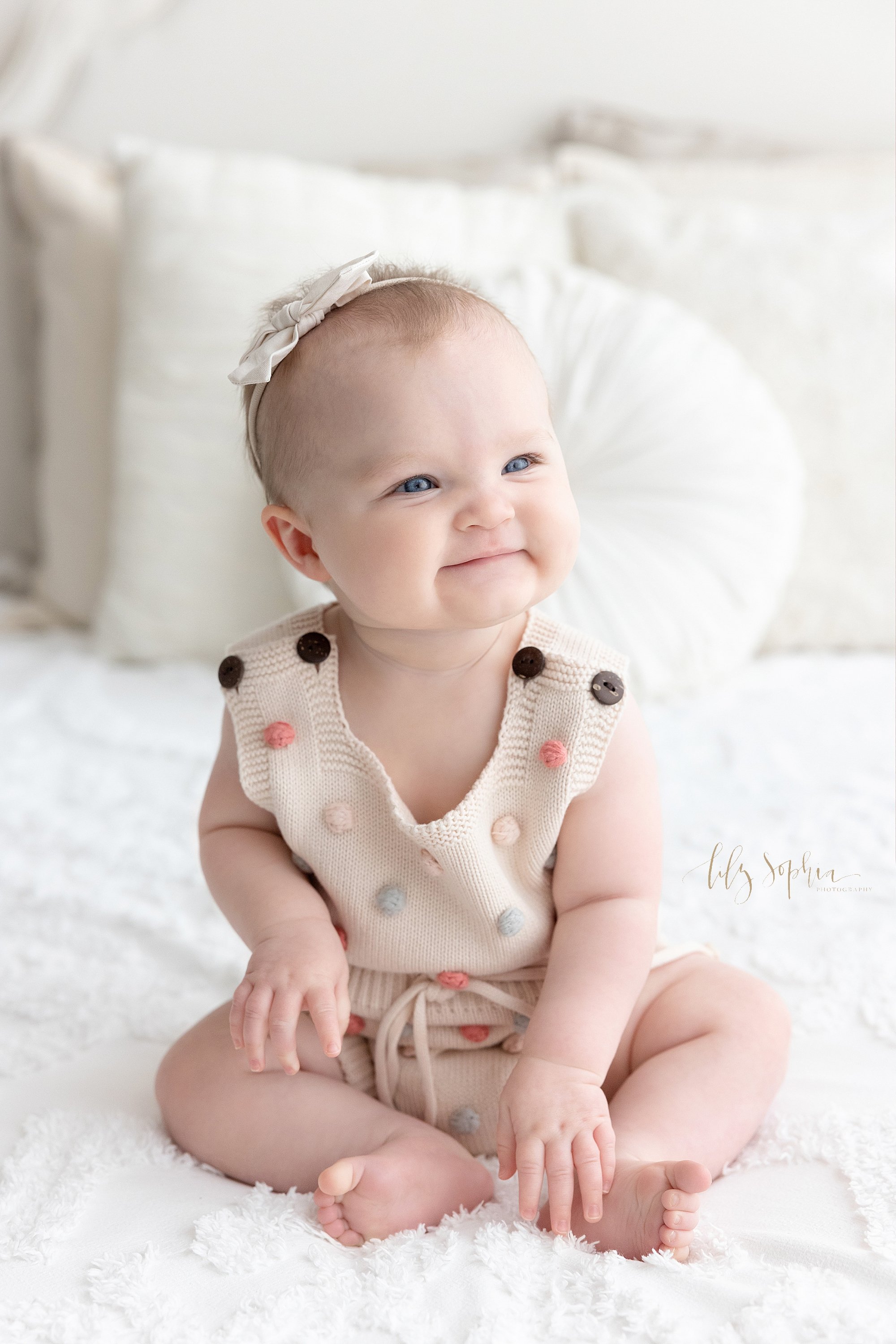  Milestone baby photo session of a smiling baby girl wearing a headband bow on her head as she sits on a bed next to a window streaming natural light in a photography studio in Ponce City Market in Atlanta. 