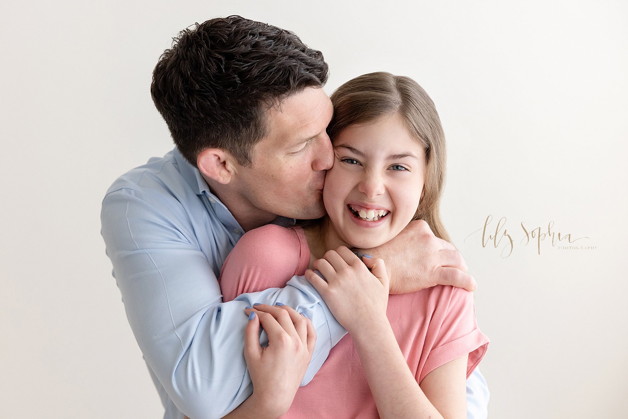  Fun family photo of a father wrapping his arms around his daughter as he stands behind her and kisses her on her cheek taken near Roswell in Atlanta in a photography studio that uses natural light. 