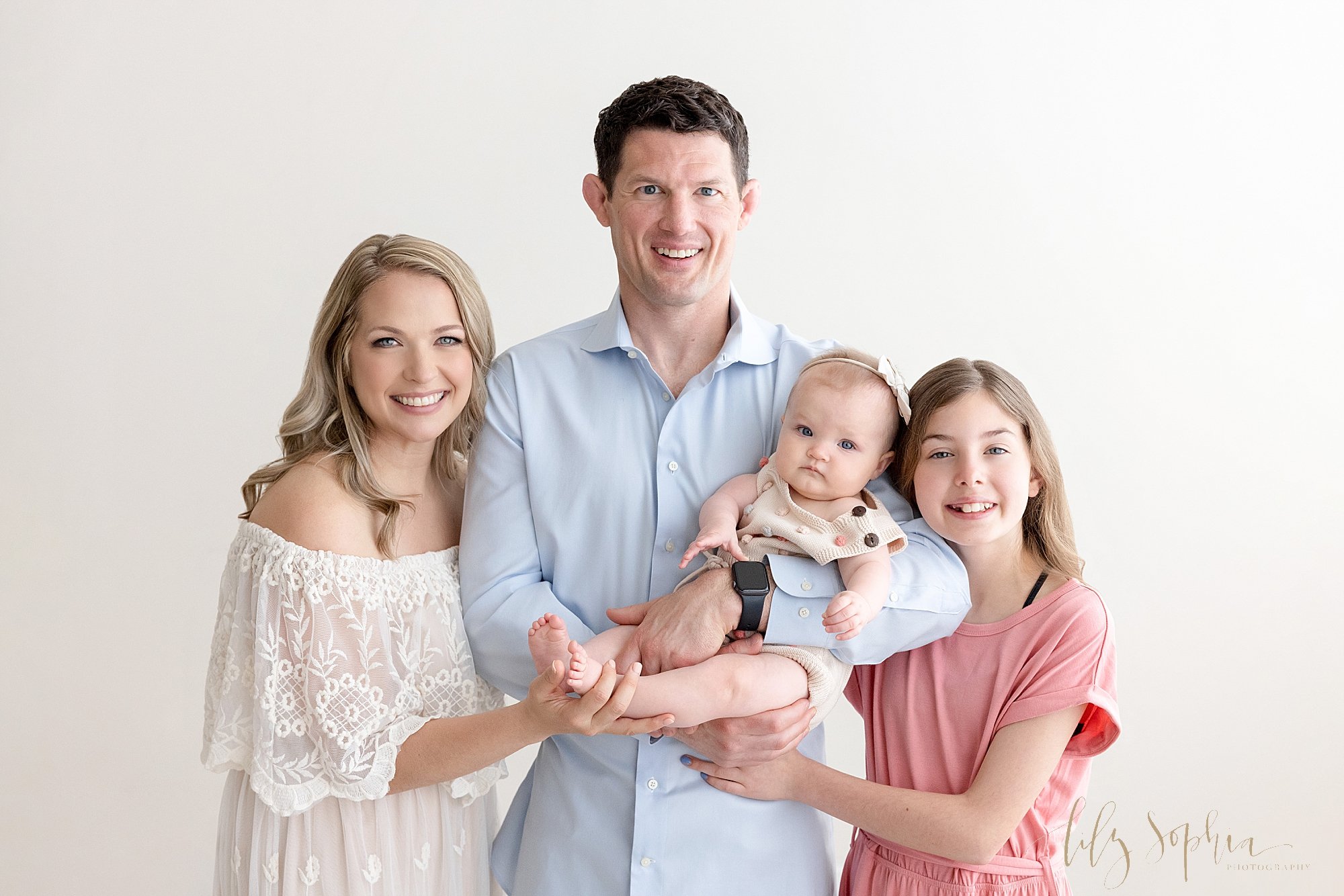  Family photo session with dad holding the baby girl in his left arm with his other daughter standing to his left while mom stands to his right and holds their daughter’s feet in her right hand taken in a natural light studio near Buckhead in Atlanta